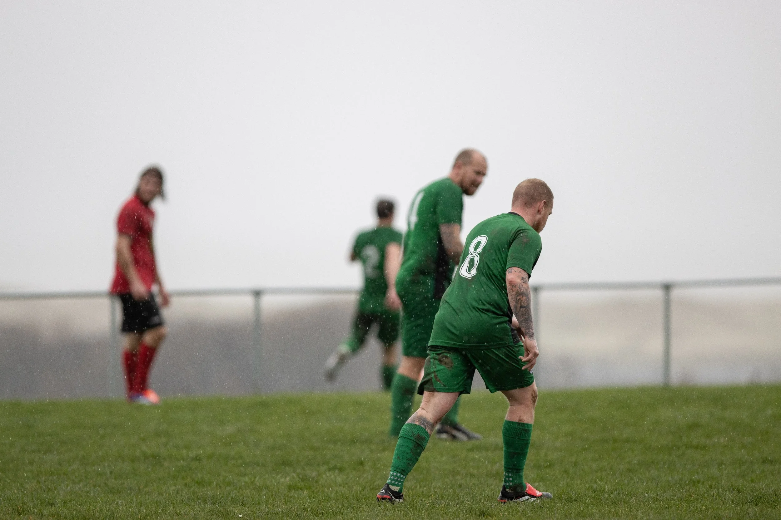 A group of soccer players on a rainy field, with three players in green jerseys and one player in a red jersey. The players appear to be disappointed or tired.
