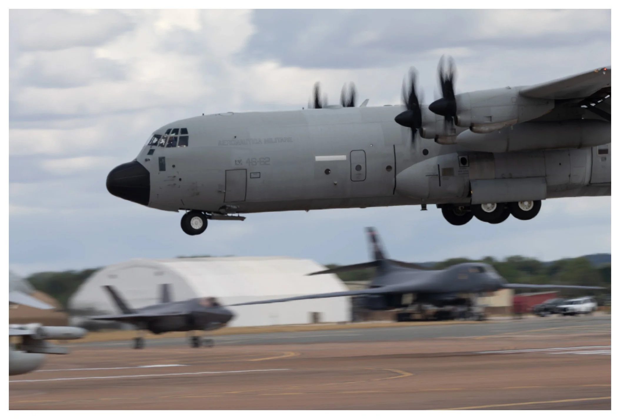 A military C-130 Hercules aircraft in flight during takeoff or landing at an airfield, with other military jets and hangars visible below on the ground.