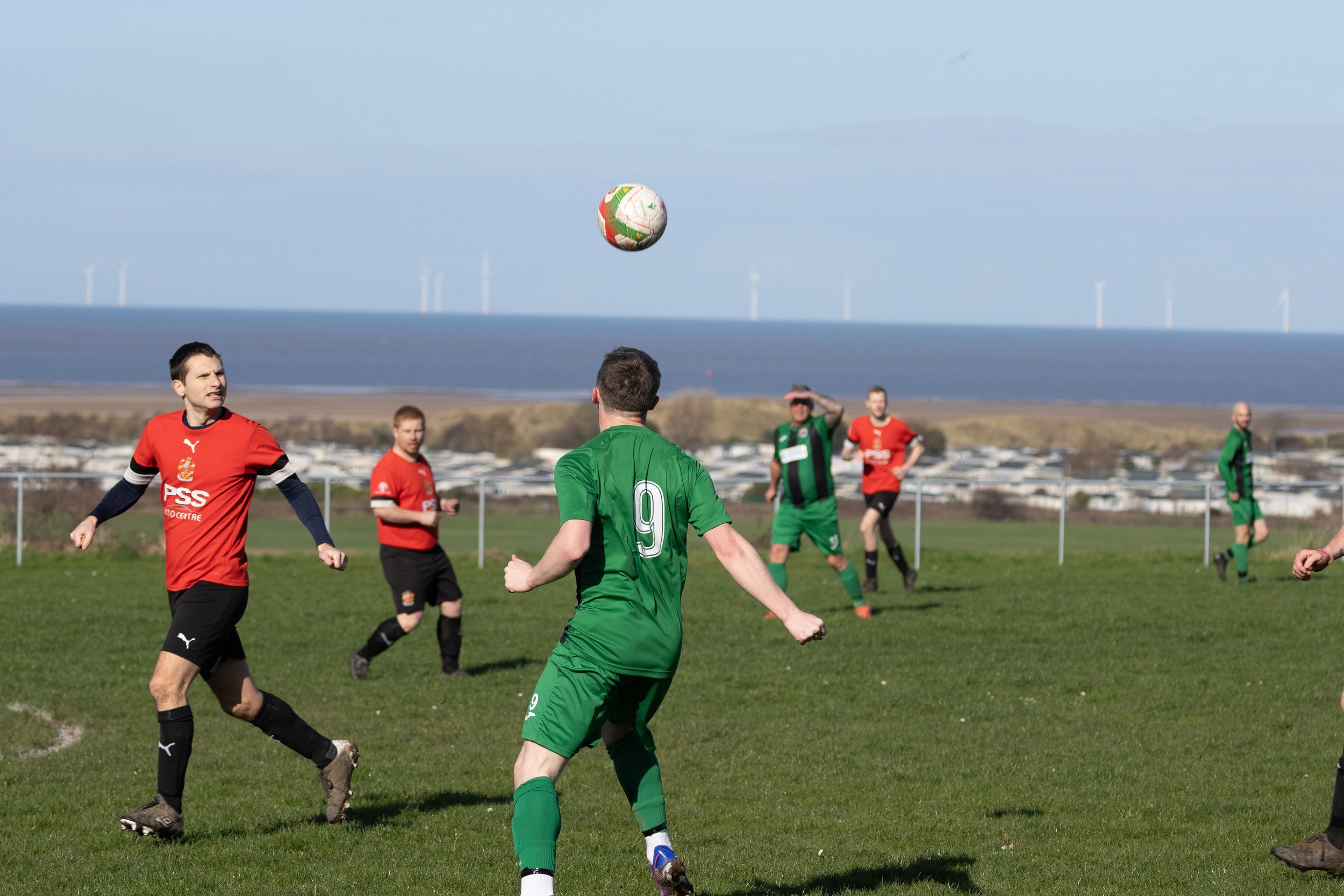 Soccer players on the field playing a game, with a ball in mid-air, and a scenic background of water and wind turbines.