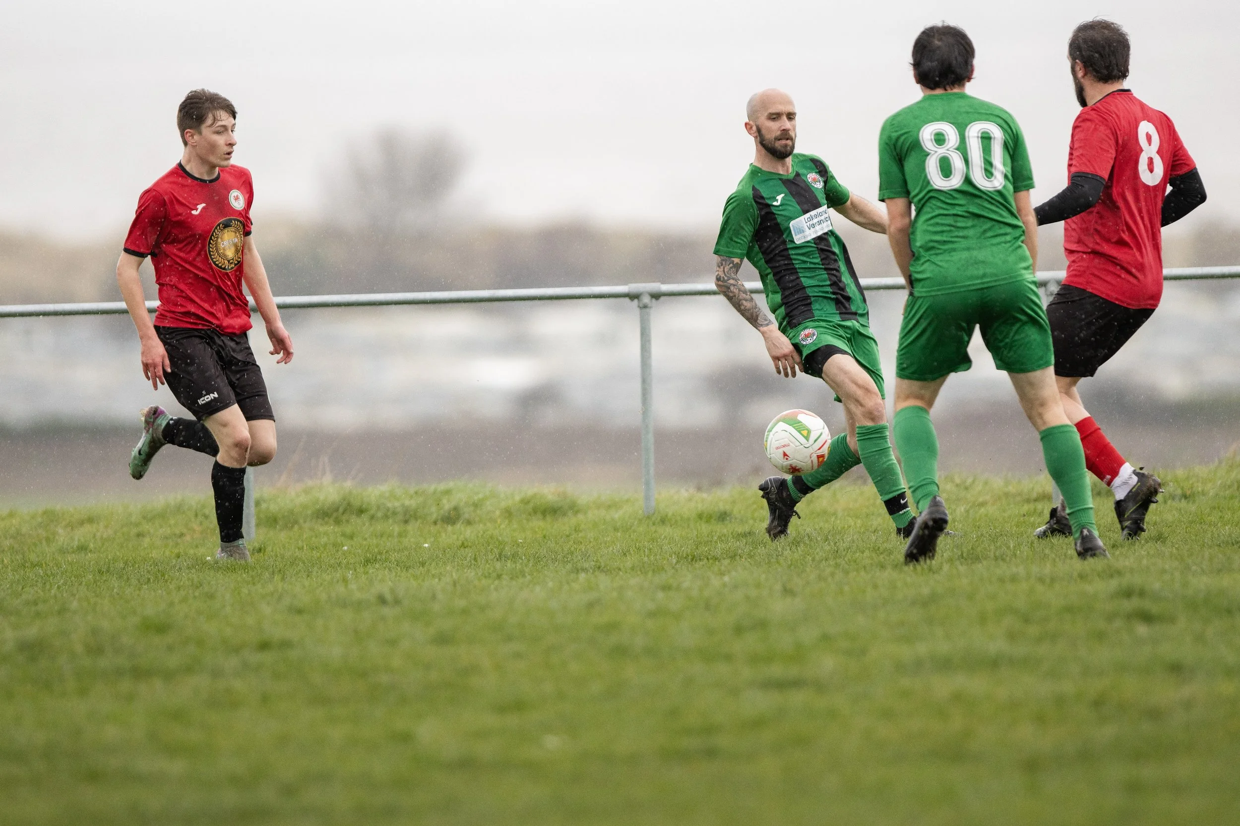 Four soccer players on a grassy field during a game, with two players in red and two players in green, competing for the ball under a cloudy sky.