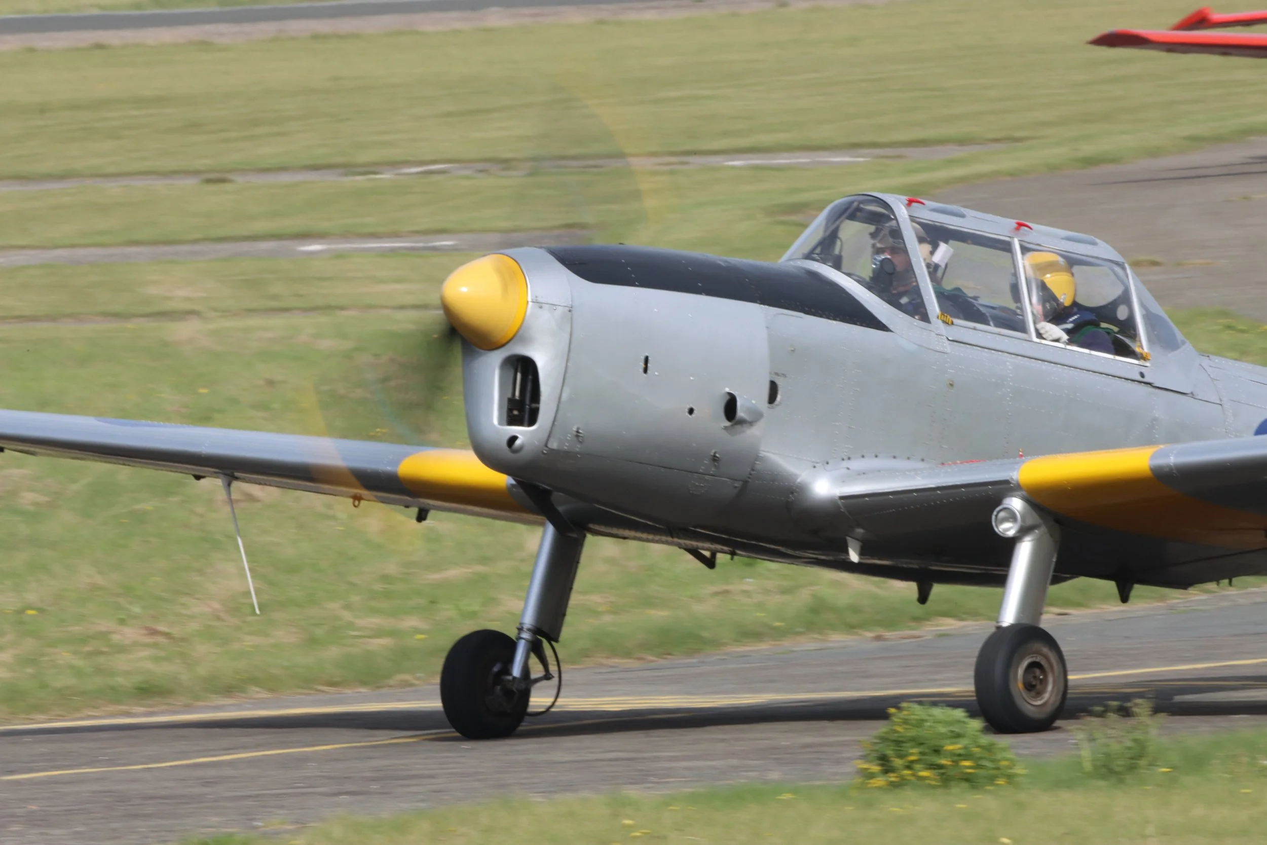 A vintage silver and yellow airplane with a glass canopy, taxiing on a runway, with two pilots inside wearing yellow helmets.