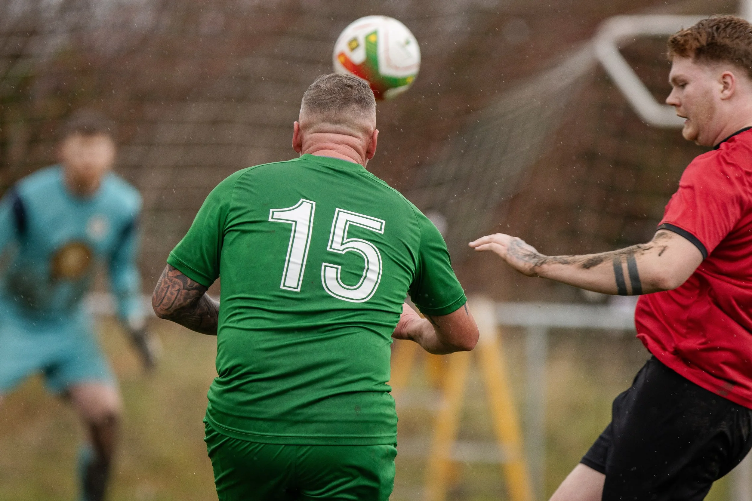 Two soccer players during a match, with one in a green jersey numbered 15 and a red jersey, both focused on a ball in mid-air, with a goalkeeper in the background.