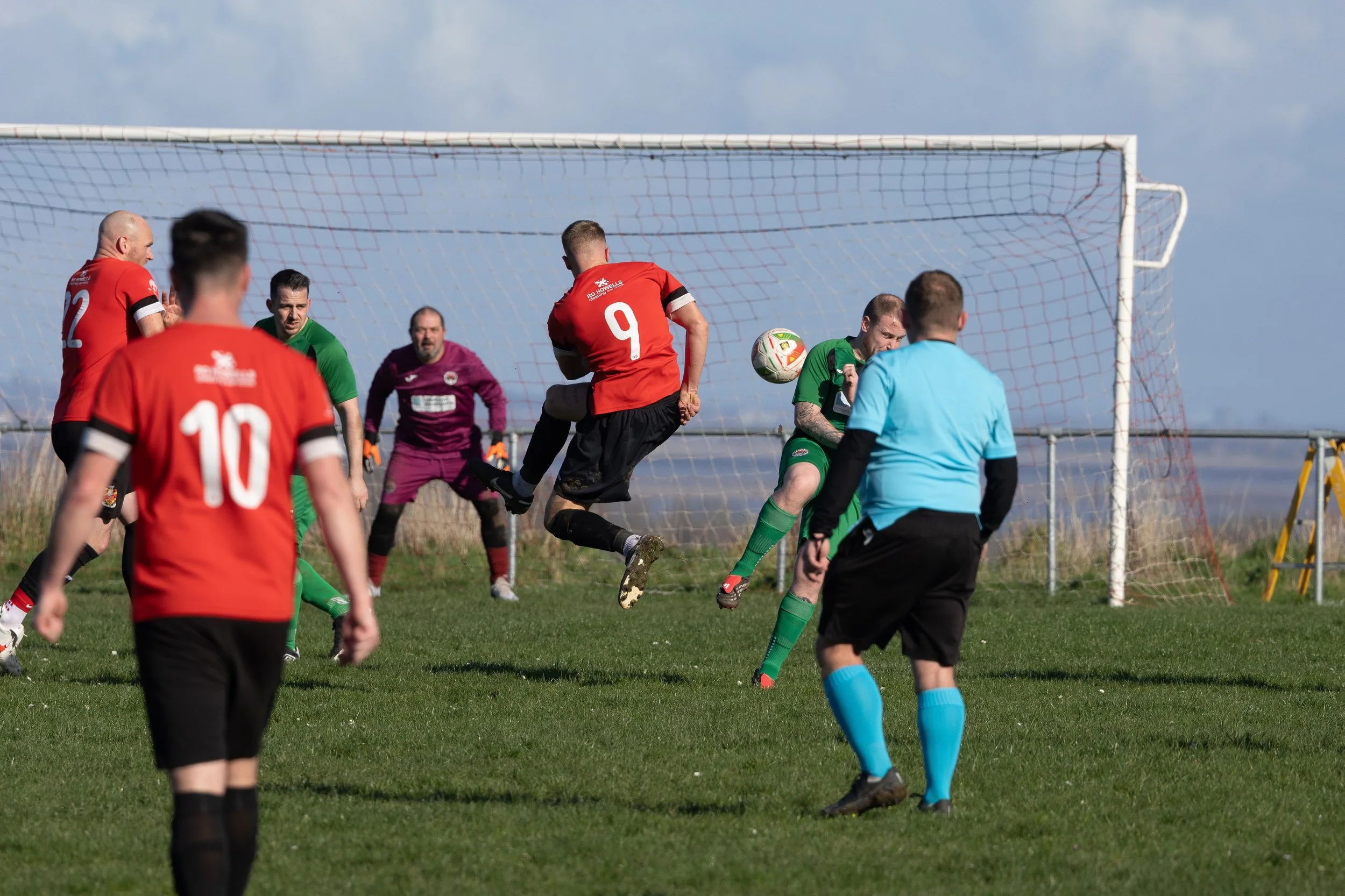 Soccer players in red and green uniforms playing near the goal, with a referee watching, during a match on a grassy field under a blue sky.
