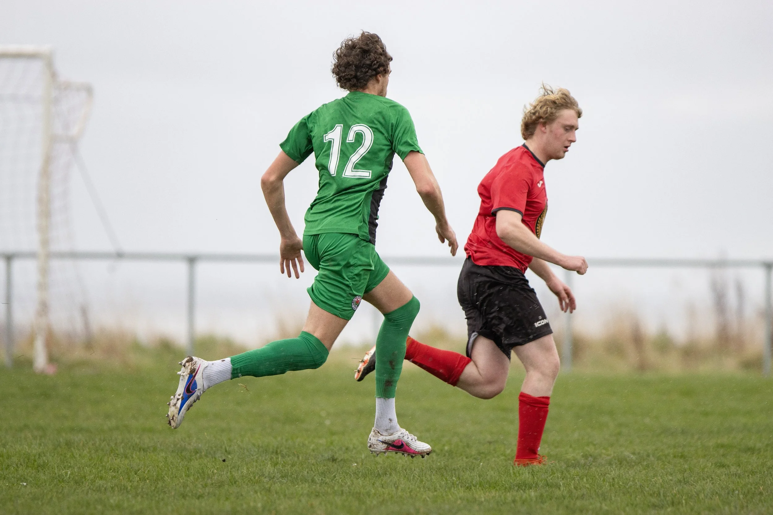 Two soccer players, one in green uniform with number 12 and one in red, running on a grassy field during a game under an overcast sky.