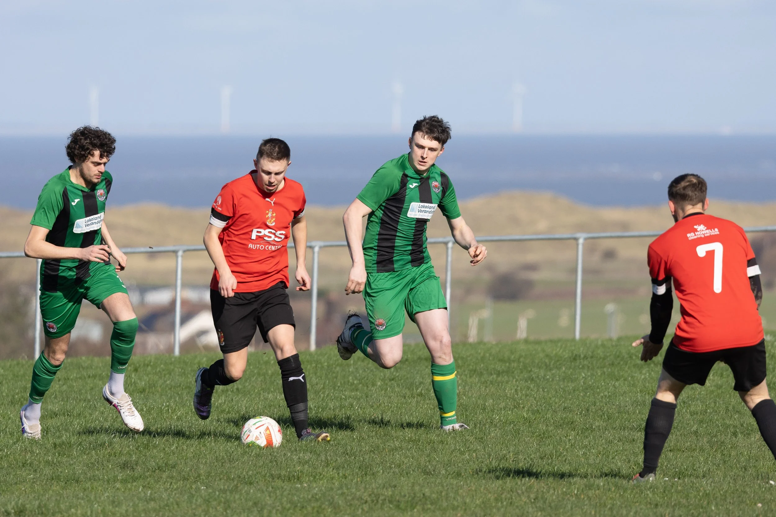 Soccer players competing for the ball on a grassy field with landscape and wind turbines in the background.