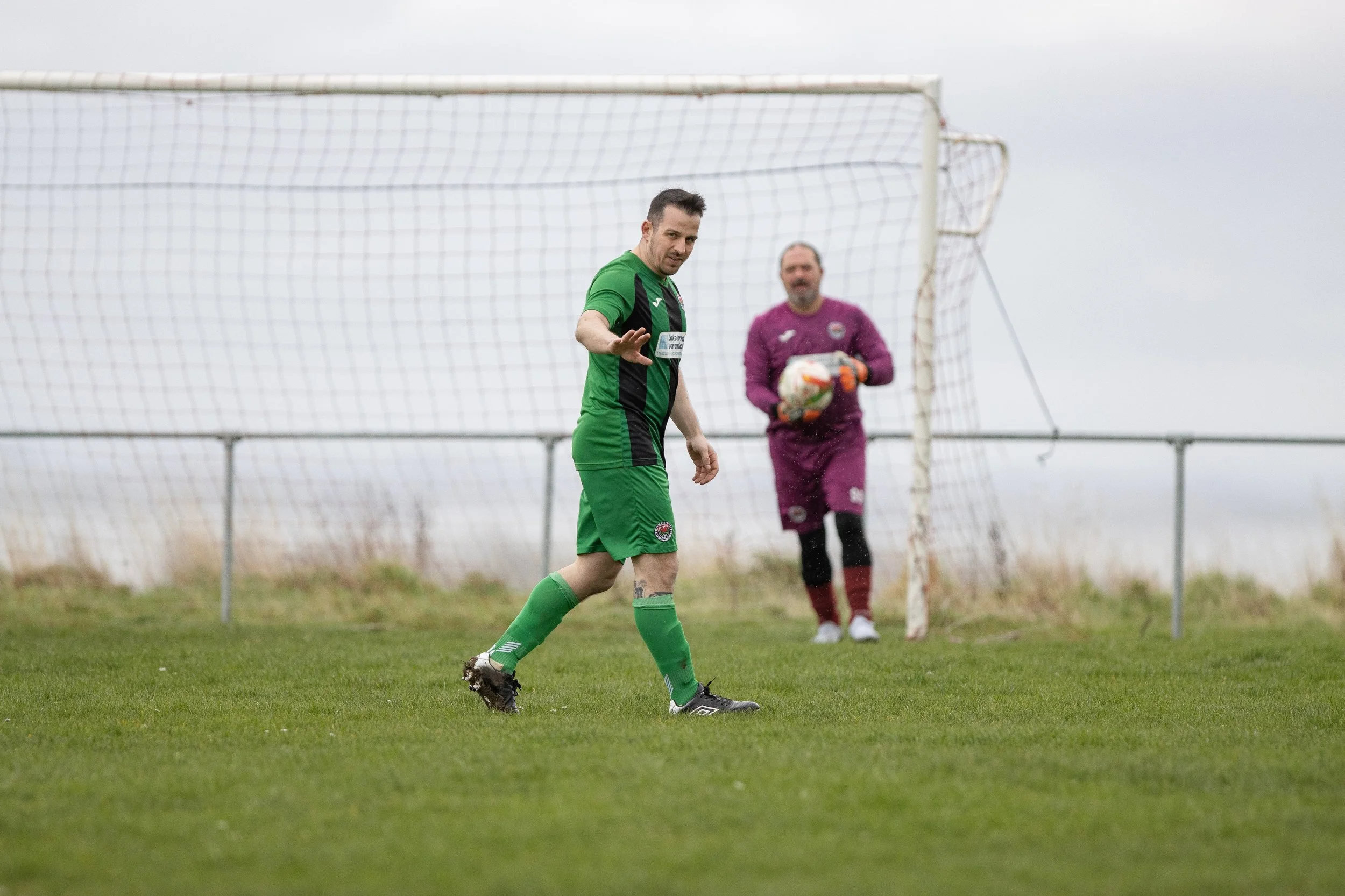 A soccer player in a green uniform waving, with a goalkeeper in a purple uniform holding a ball in the background, on a grassy field near the goal.