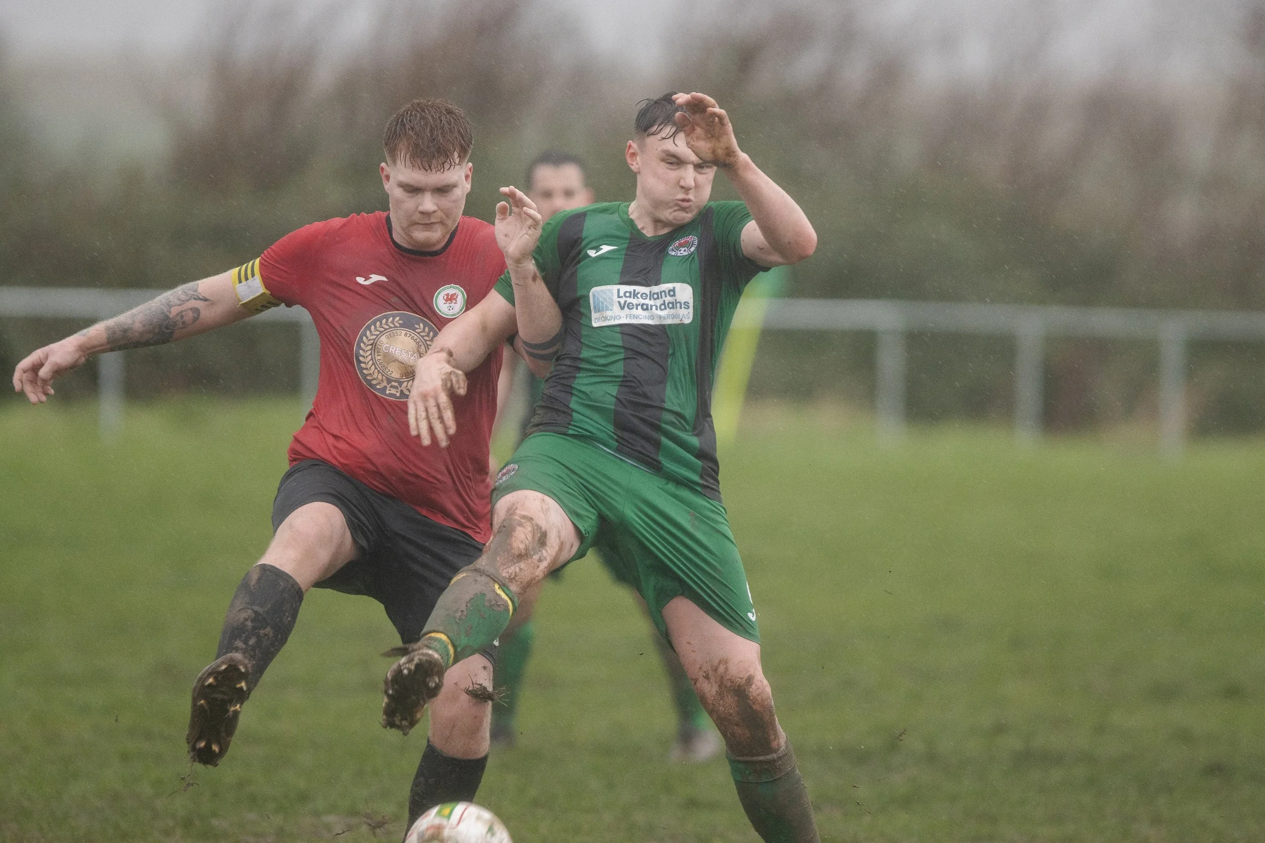 Two soccer players competing for the ball on a muddy field in rainy weather, one wearing a red jersey and the other in a green and black jersey.