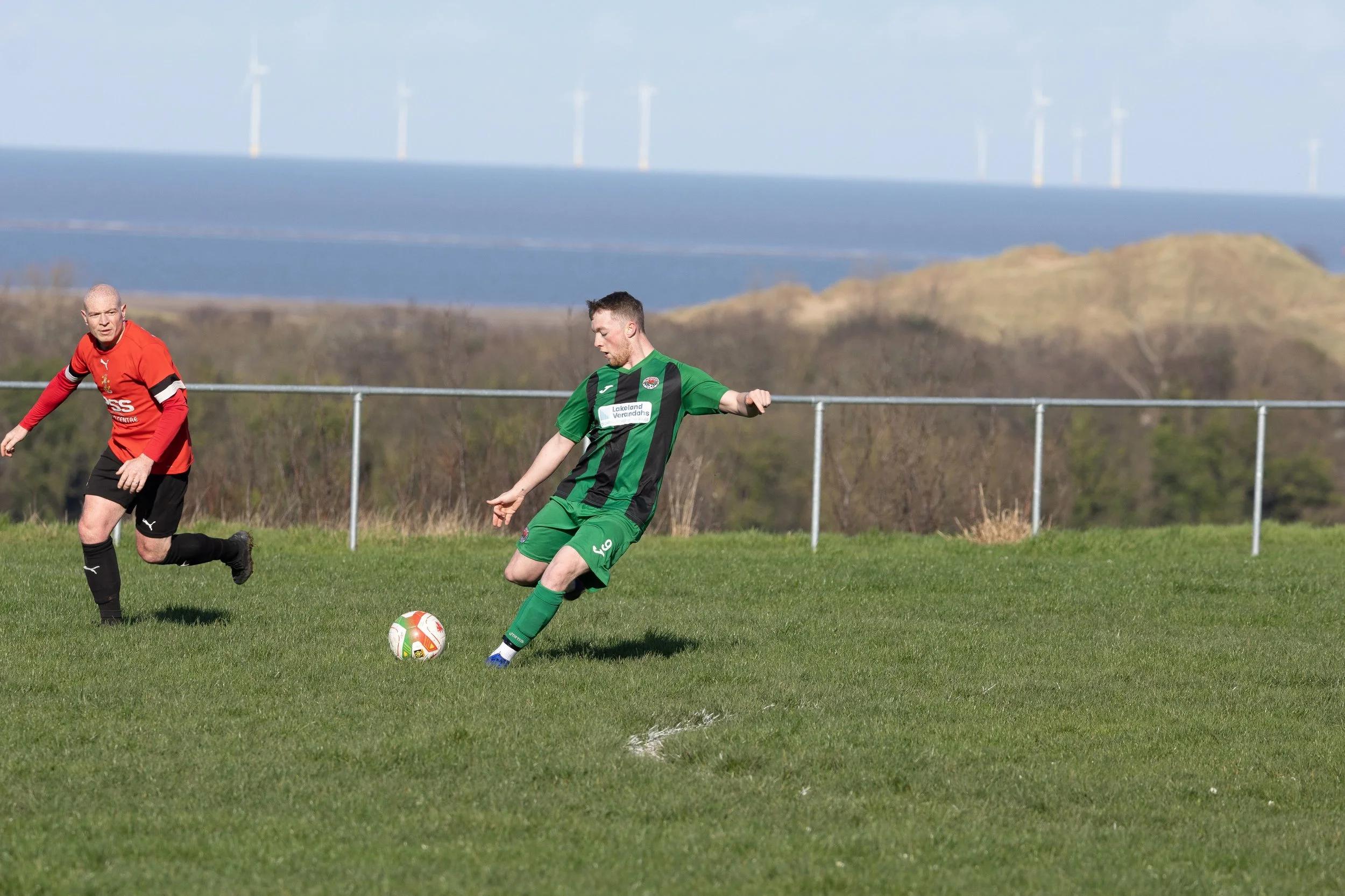 Two men playing soccer on a grassy field with hills and wind turbines in the background. One man is in a red jersey and black shorts, and the other in a green and black jersey with green shorts, about to kick the ball.