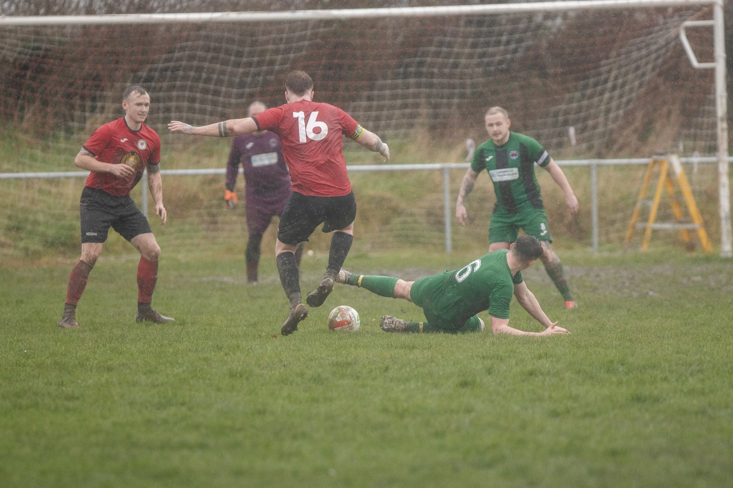 Soccer players in green and red jerseys competing for the ball on a muddy field during a match in rainy weather.