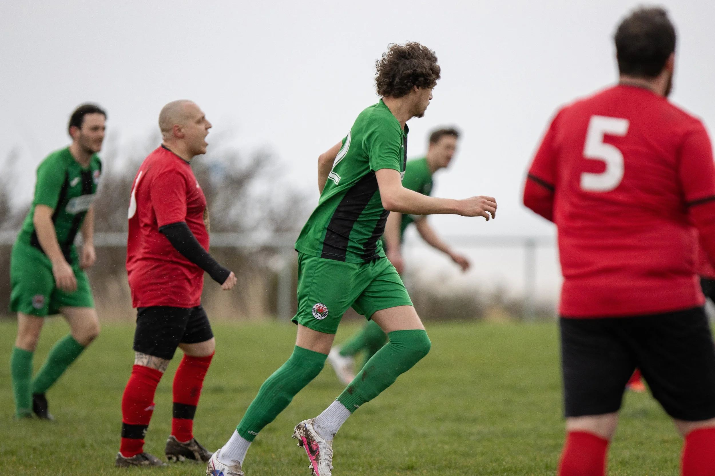 Soccer players in green and red uniforms on a grassy field during a match, with overcast sky in the background.