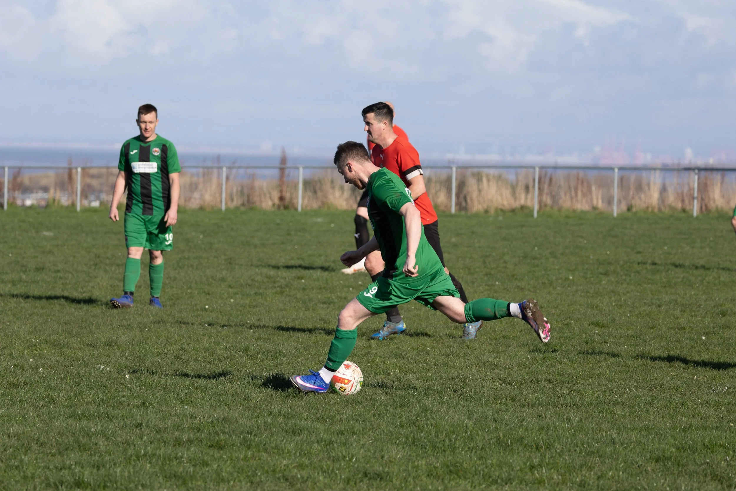 A soccer player in a green uniform is kicking a ball on a grassy field, with two other players in red and black uniforms nearby, and a player in green watching from a distance.