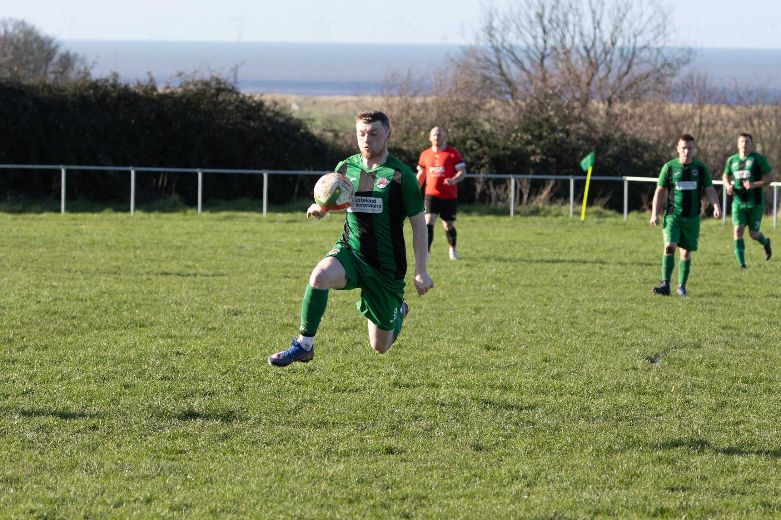 A soccer player in a green and black uniform is jumping to control the ball on a grassy field during a match, with other players and trees in the background.