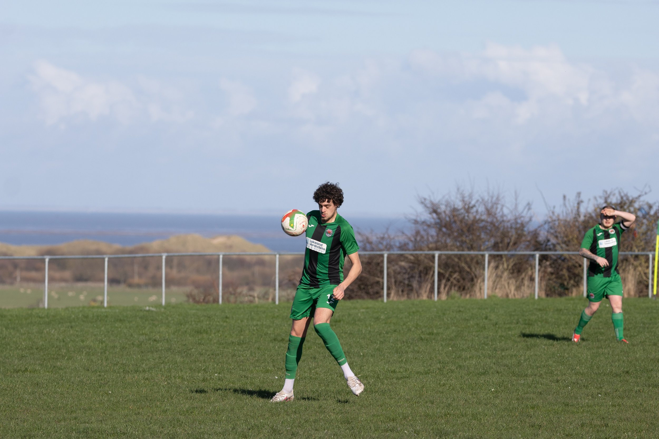 Two soccer players in green uniforms on a field, one controlling the ball with his foot, with a landscape and water in the background.