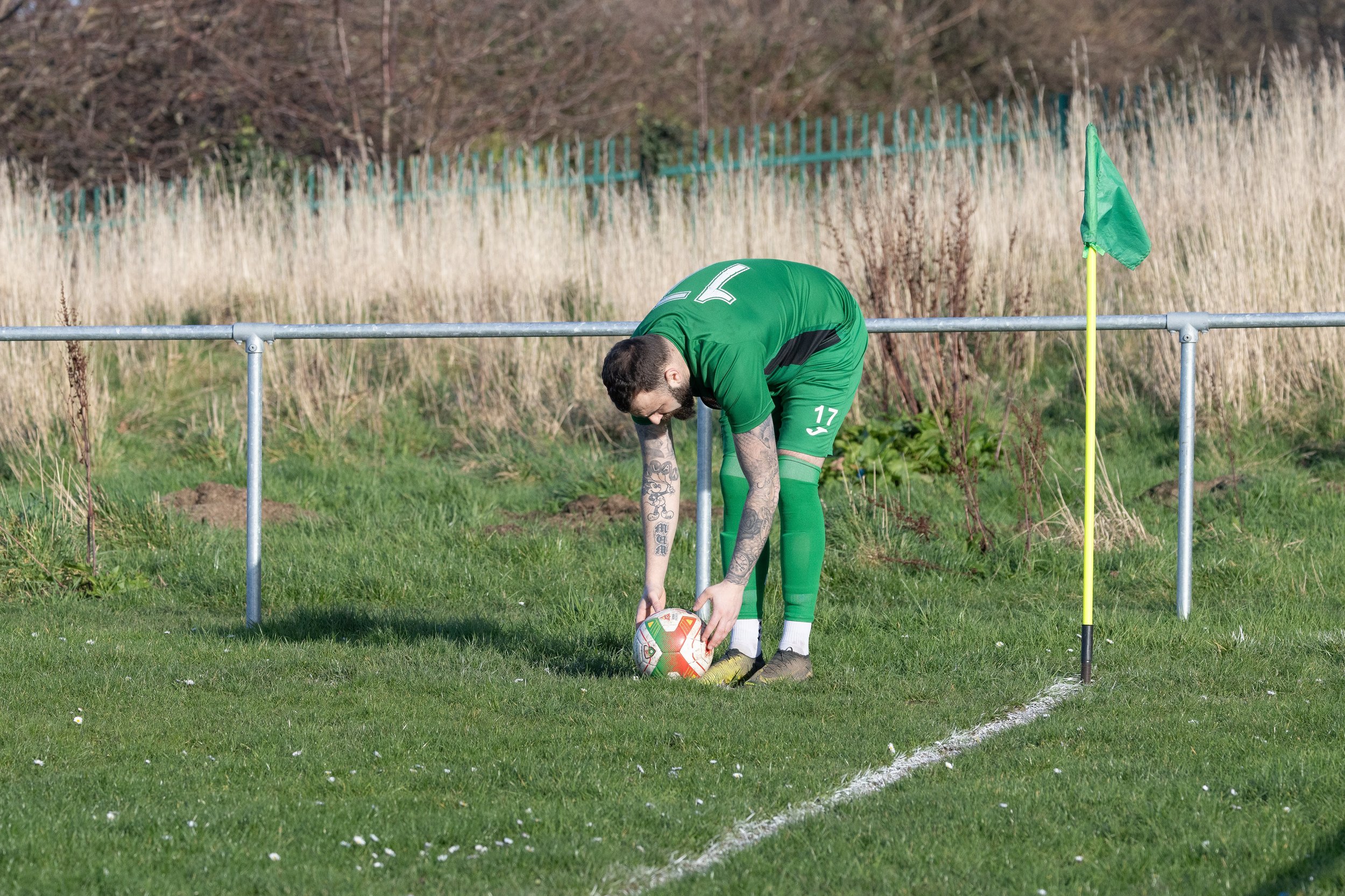 A soccer player wearing a green uniform with the number 17 on his shorts is bending down on the field, placing the ball at the corner of the field near the corner flag.