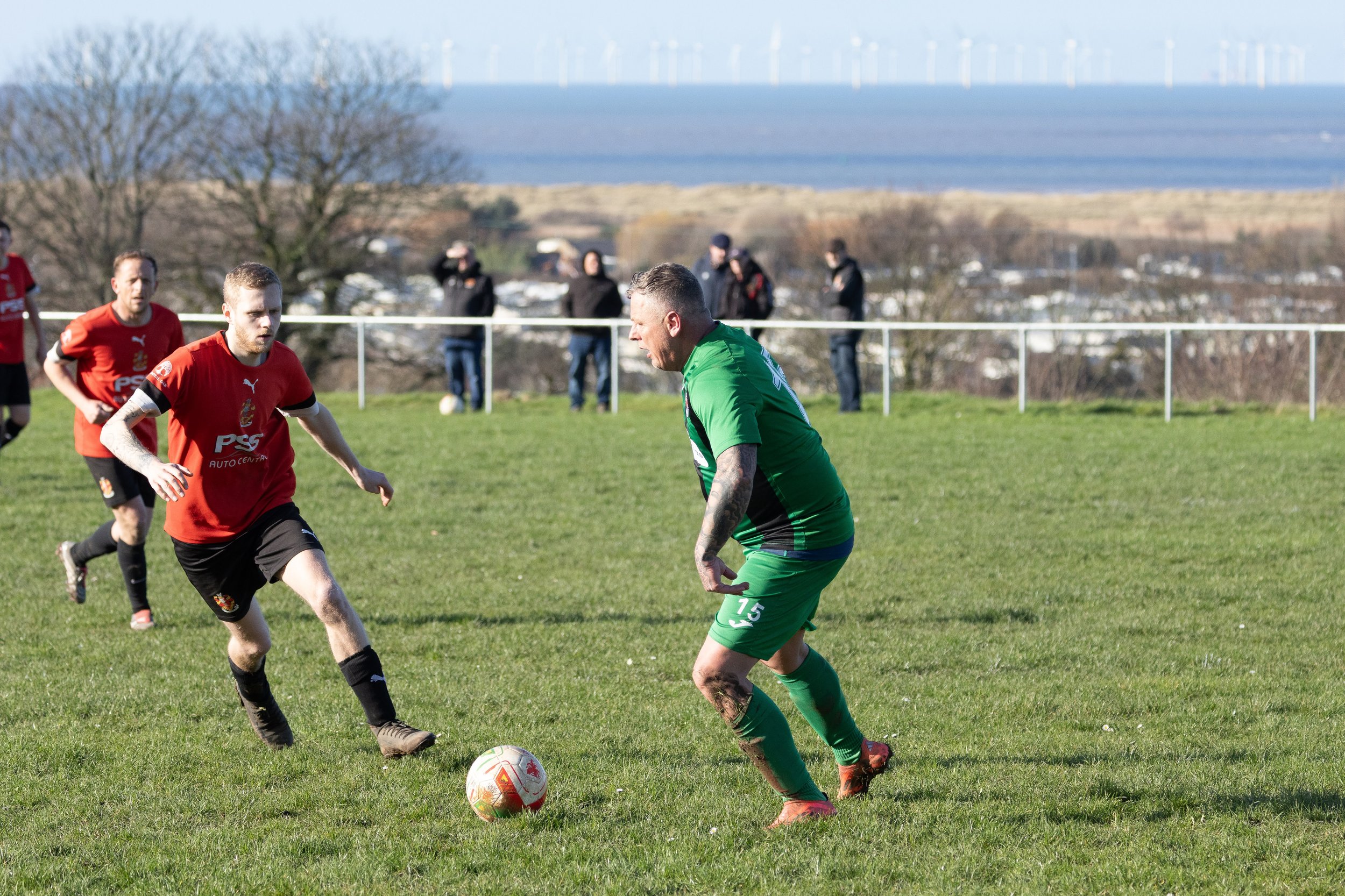 Soccer game with players in green and red jerseys on a grassy field near water and trees in the background.