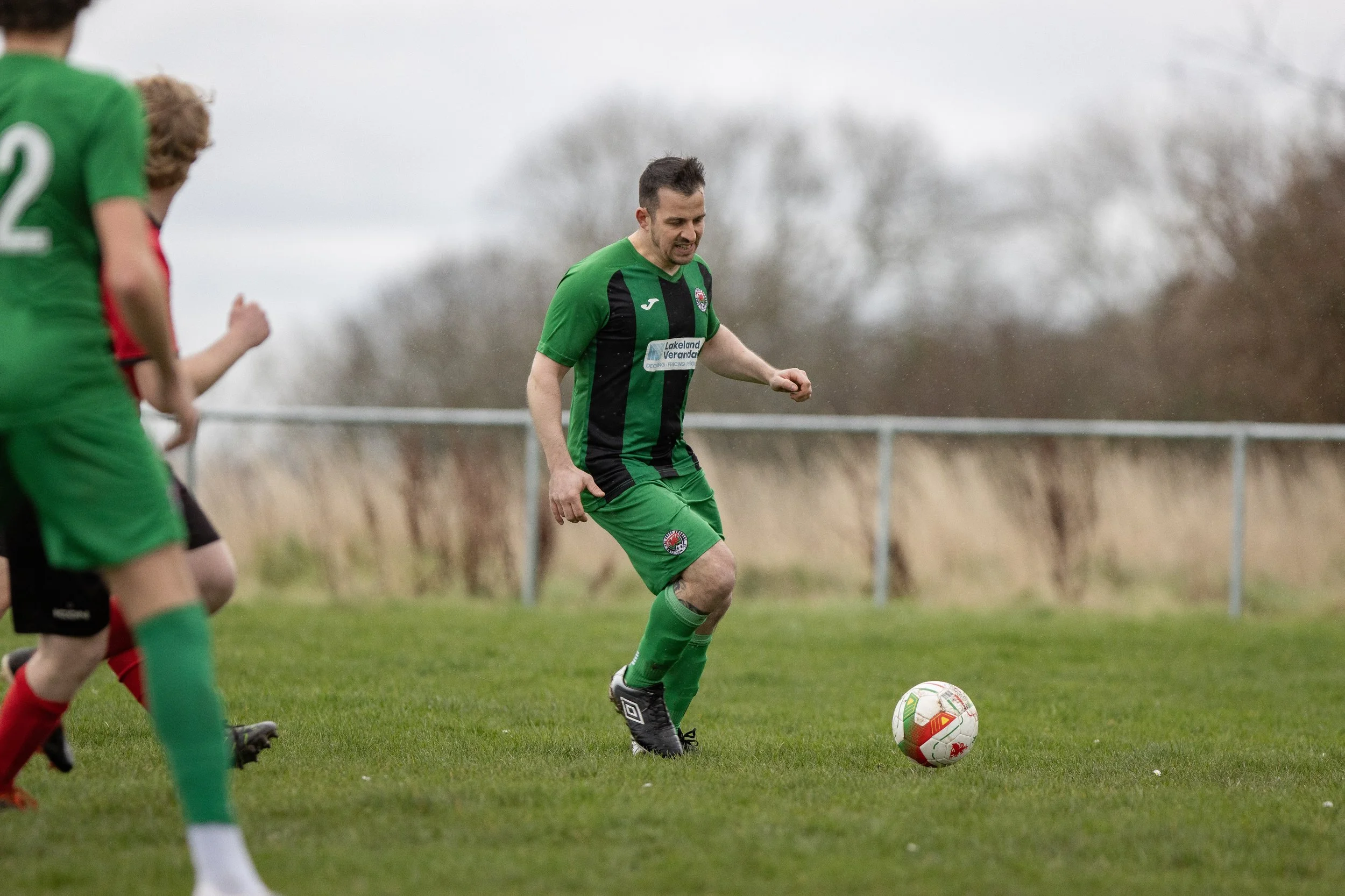 A soccer player in a green and black uniform about to kick a white soccer ball on a grassy field, with other players partially visible nearby.