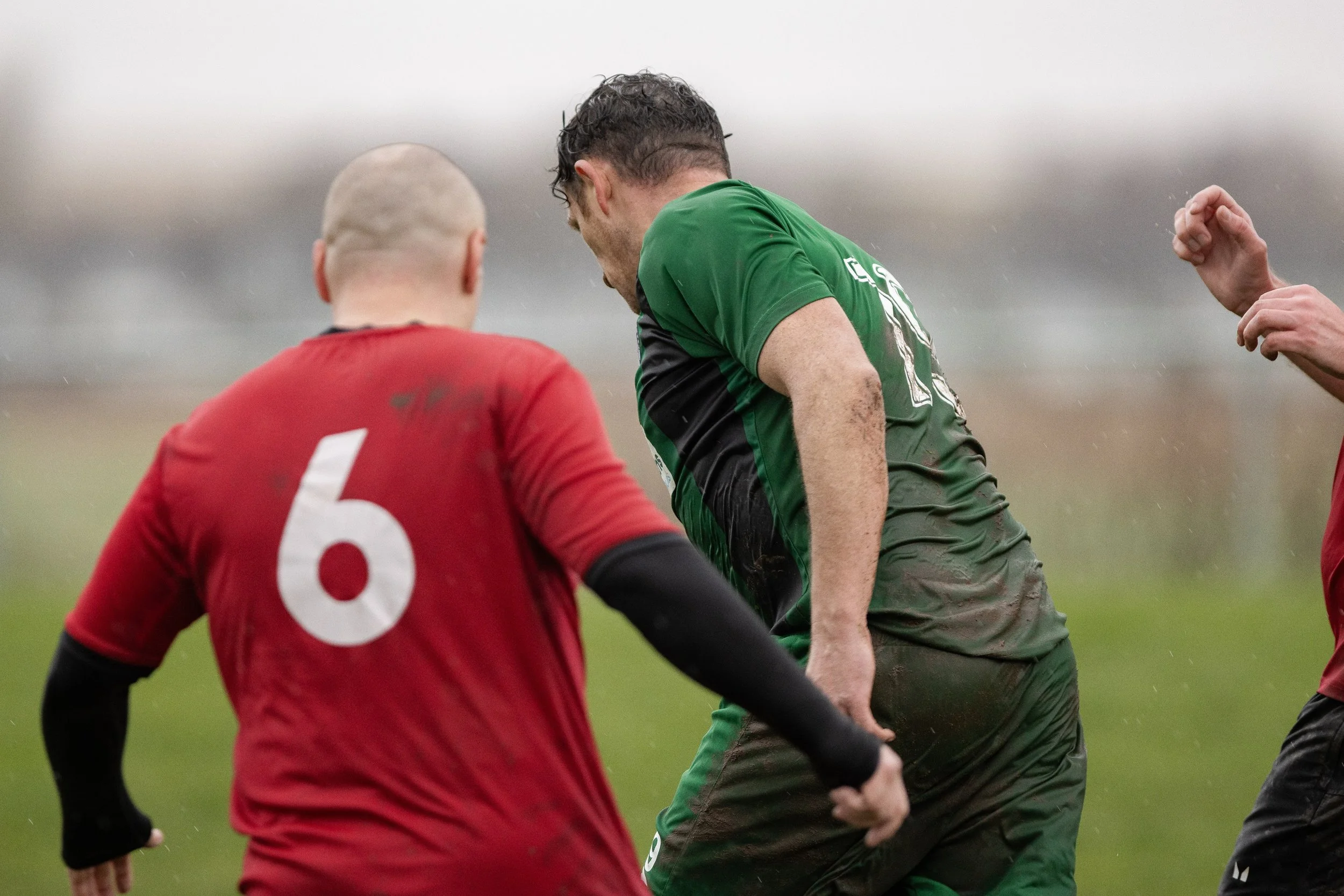 Two soccer players, one in a red jersey with the number 6 and one in a green jersey, on a muddy field during a match under rainy conditions.