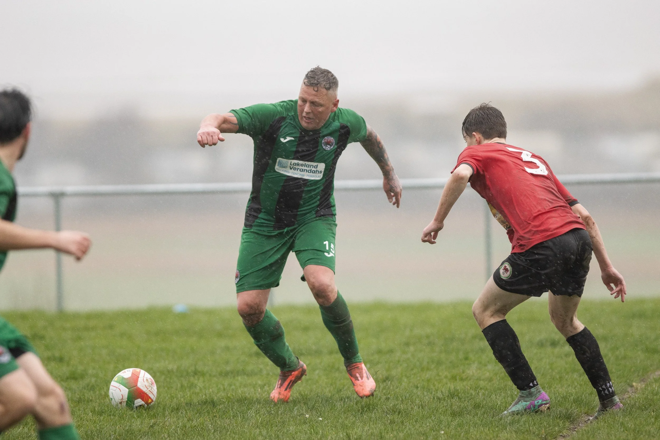 Soccer players in rain, one in green and one in red, competing for the ball on a grassy field.