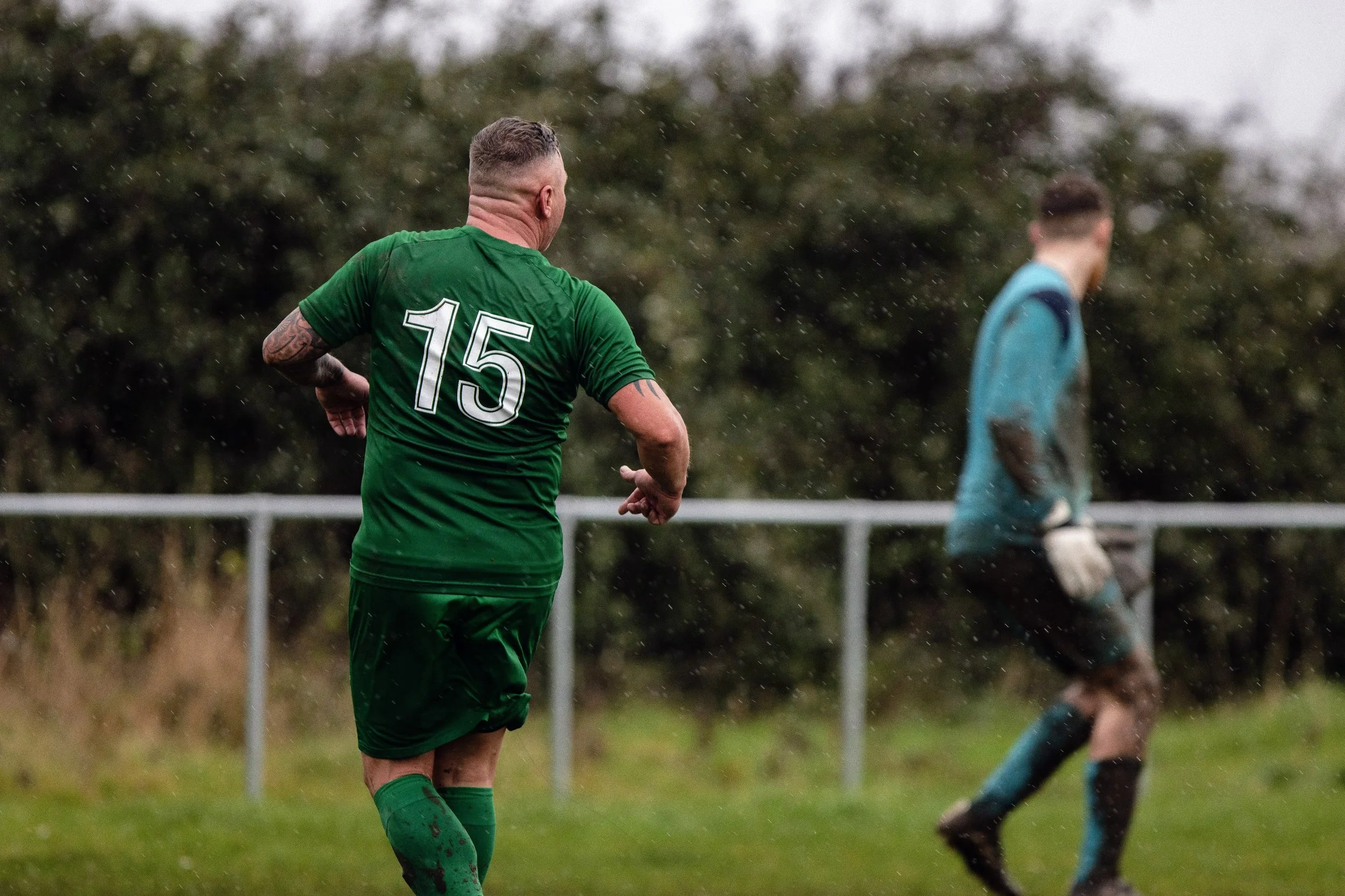 A soccer player wearing a green uniform with the number 15 on his back, seen from behind, running on a rainy field with a blurred goalkeeper in a blue and black uniform in the background.
