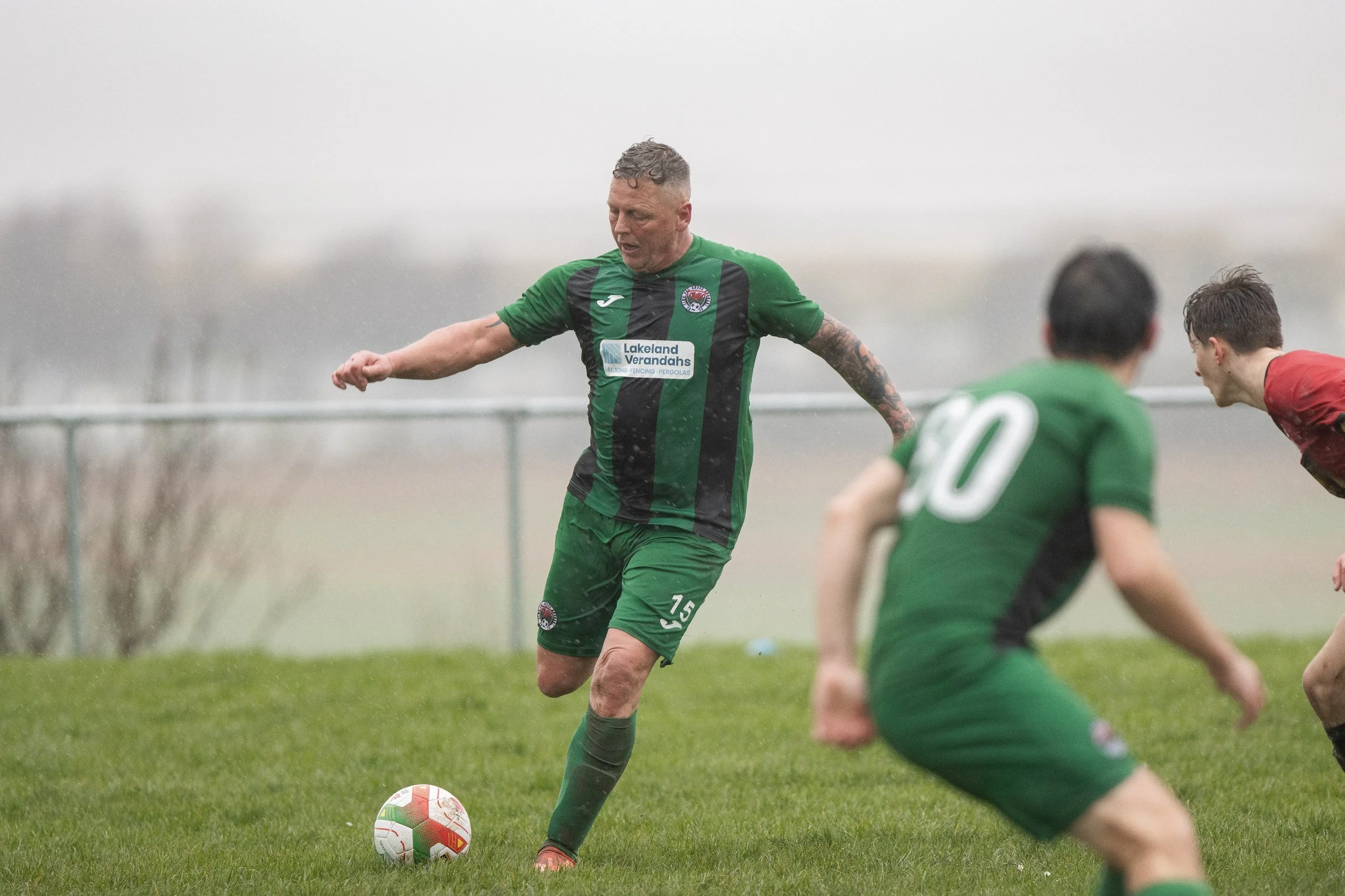 A soccer player in a green and black uniform kicking a soccer ball on a grassy field during a rainy day, with two other players nearby.