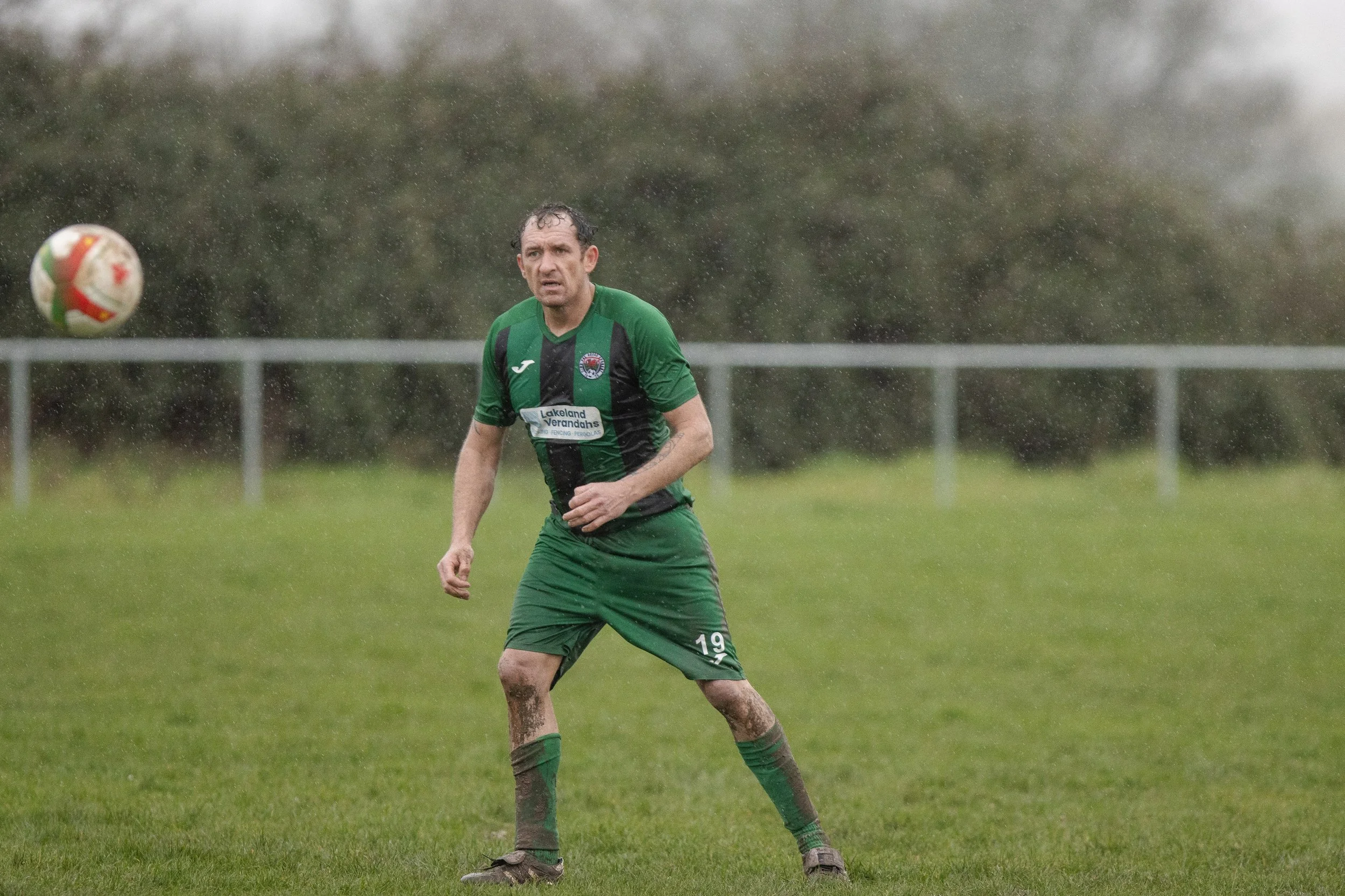 A male soccer player in green and black uniform stands on a muddy field during a rainy game, with a soccer ball in mid-air nearby.