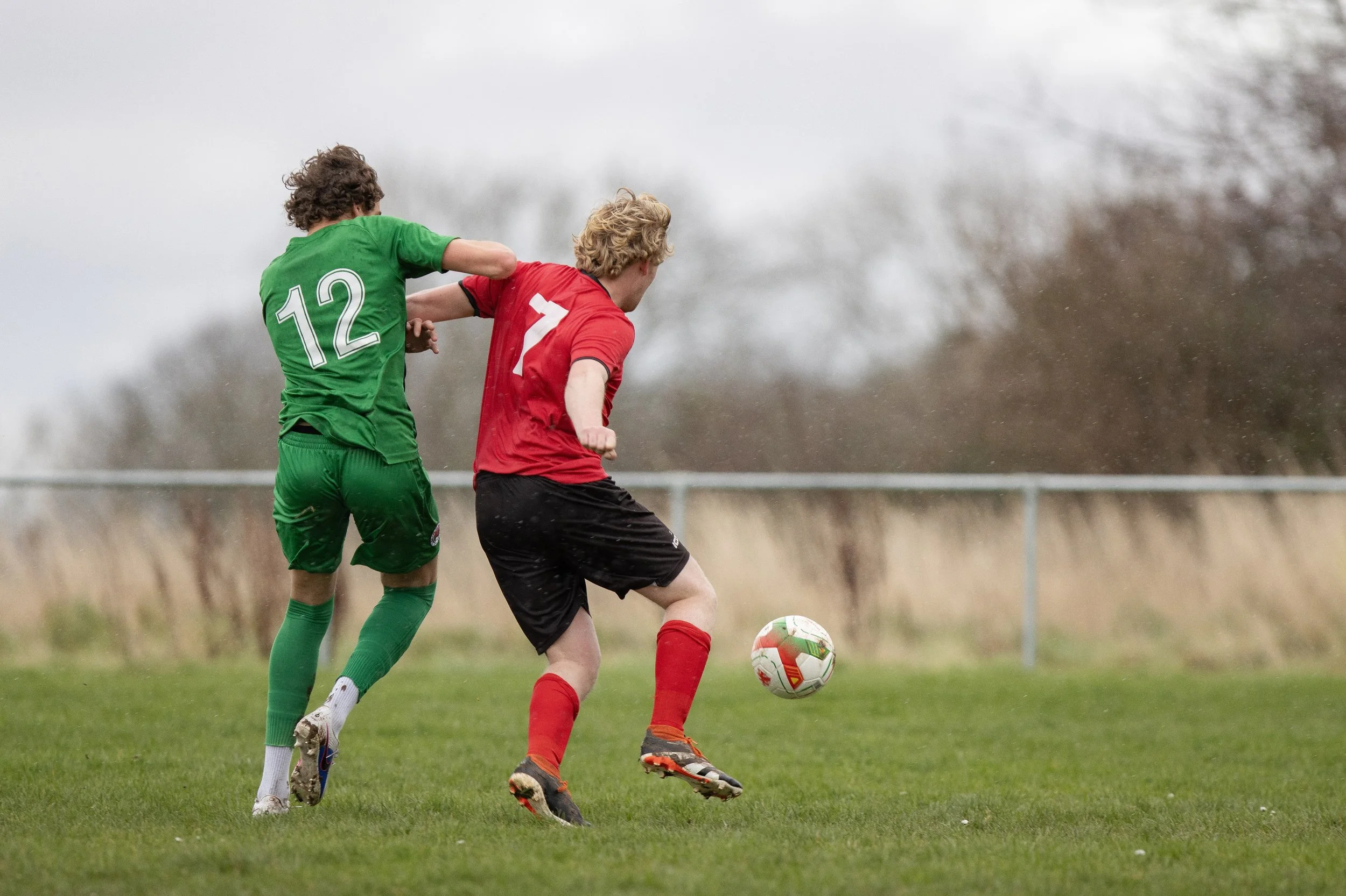 Two soccer players, one in a green jersey with the number 12 and the other in a red jersey with the number 7, are competing for the ball on a grassy field.