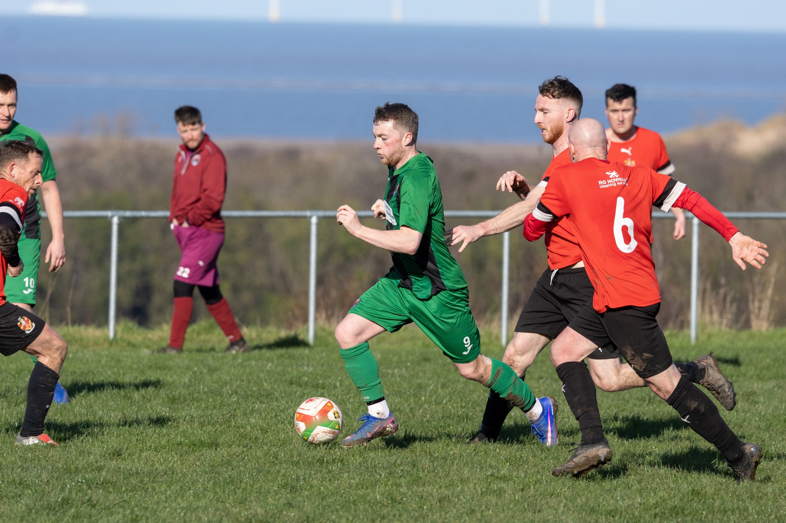 Soccer players competing for the ball on a grassy field with a fence and landscape in the background.