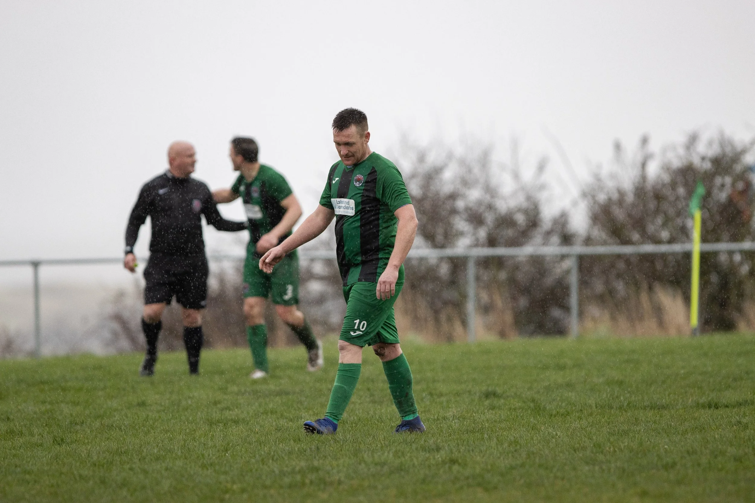 A soccer player wearing a green uniform with the number 10 is walking on a grass field, looking down with a disappointed expression. In the background, a referee and another player in a green uniform are celebrating.
