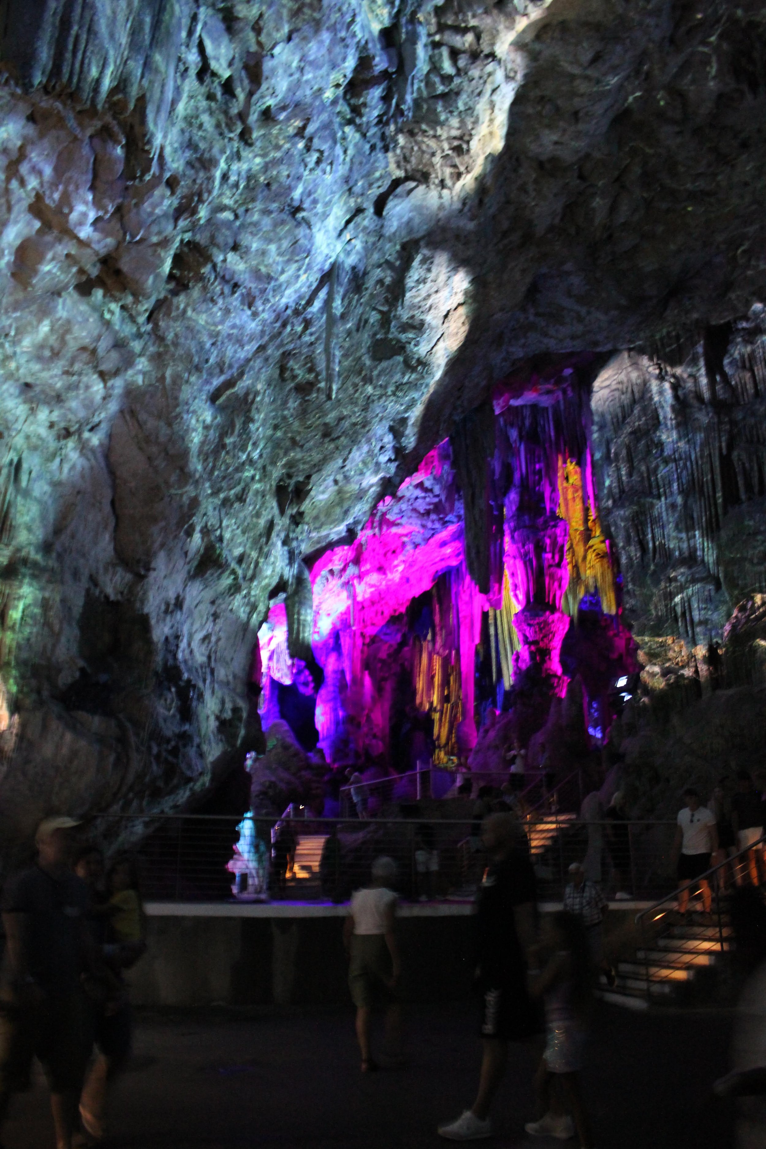 People visiting a large illuminated cave with colorful stalactites and stalagmites.