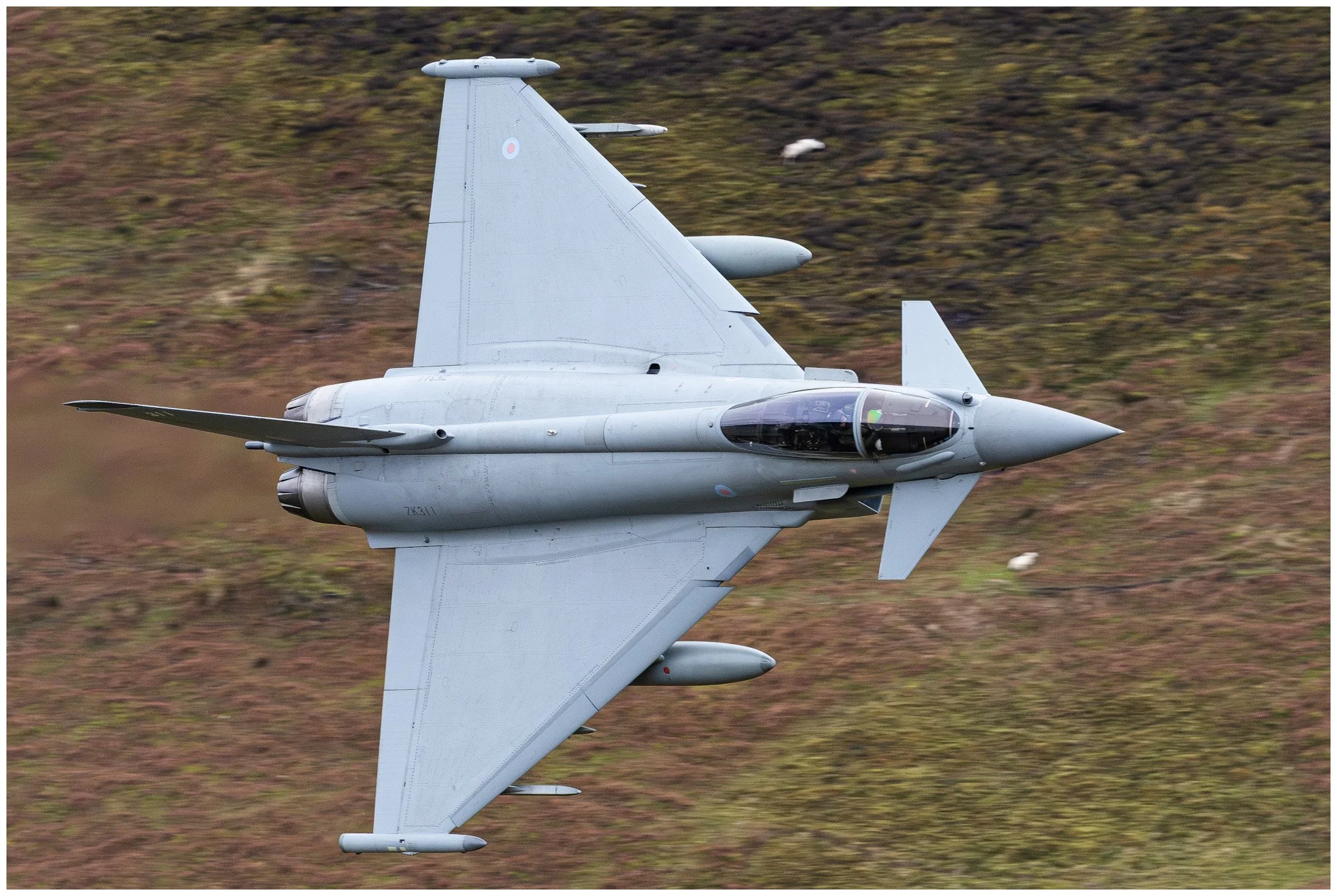 A military fighter jet flying low over a grassy hillside.