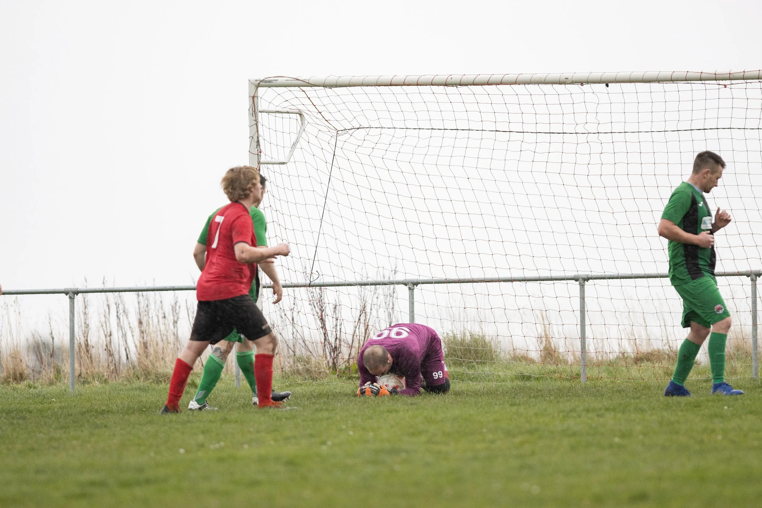 A soccer game in progress with players near the goal post. A goalkeeper in purple kneels on the ground, holding the ball, while two players in green jerseys and one player in a red jersey are nearby.