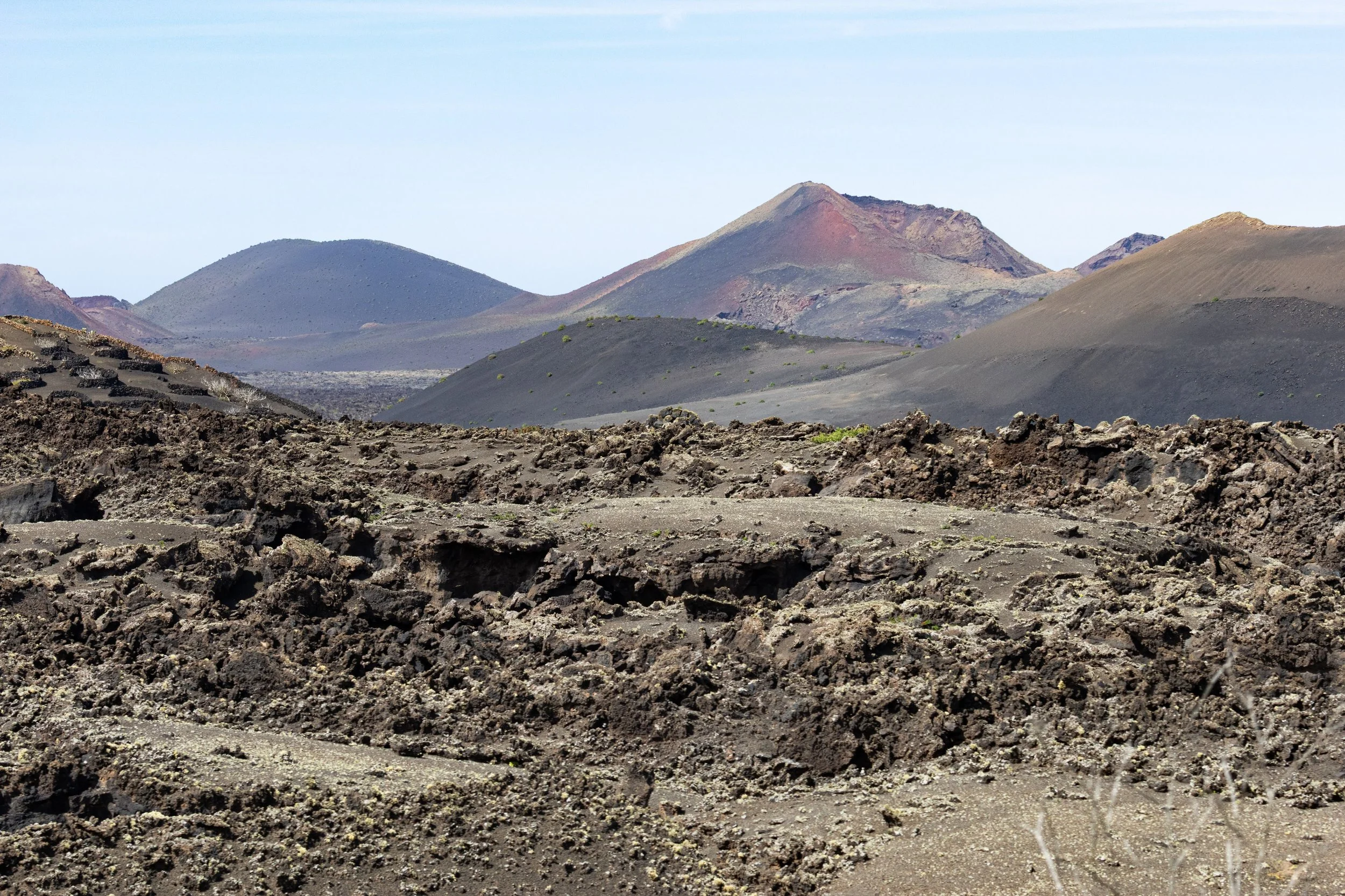 Volcanic landscape with dark rocky terrain and multiple volcanic cones in the background under a clear sky.