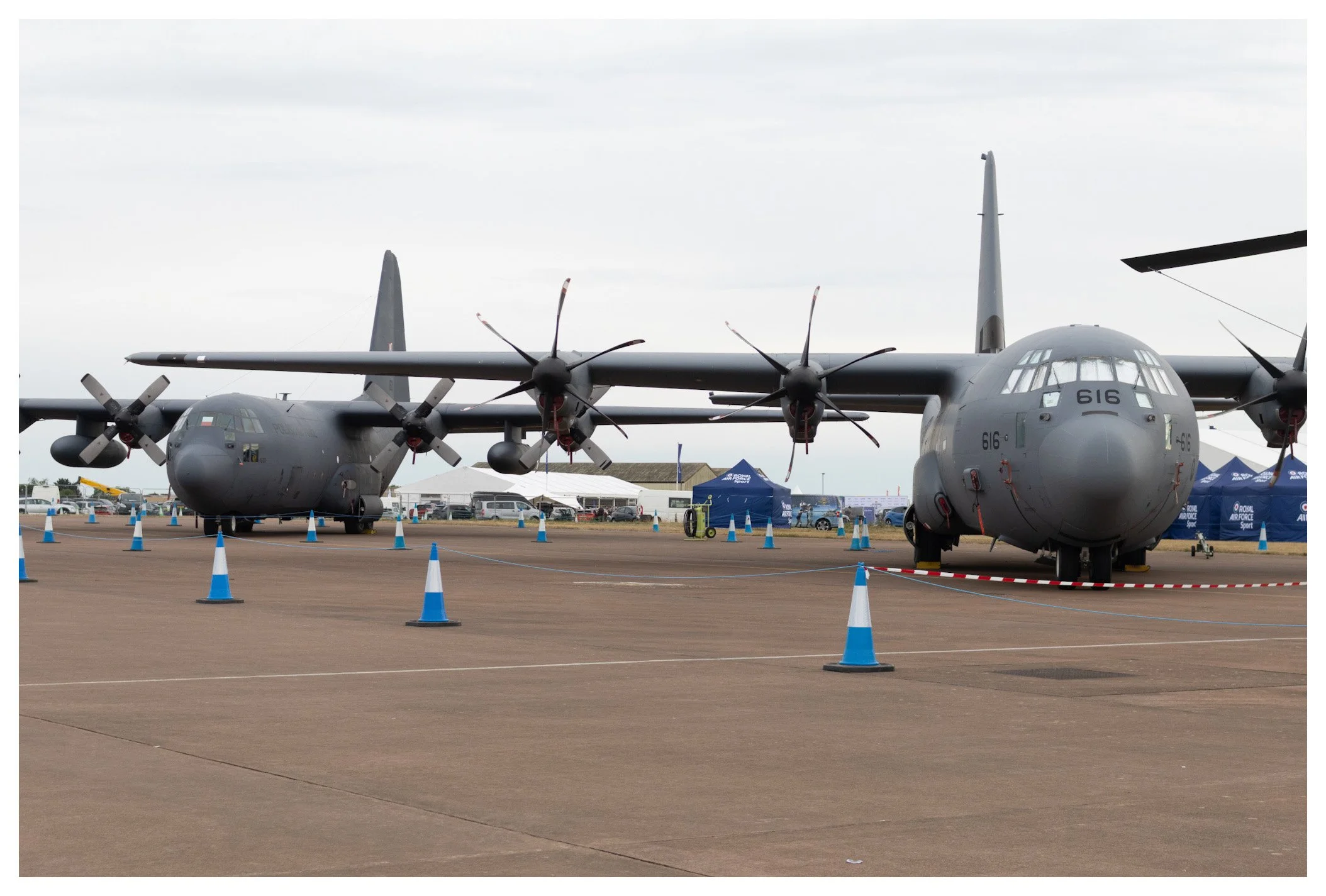 A military cargo aircraft with four propeller engines on display at an airshow, surrounded by blue cone barriers, with tents and other aircraft in the background.