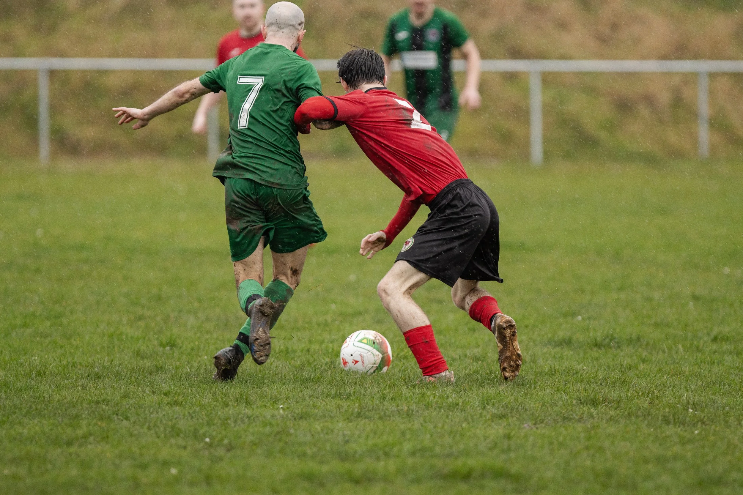 Two soccer players competing for the ball on a muddy field during a rainstorm, with additional players in the background.