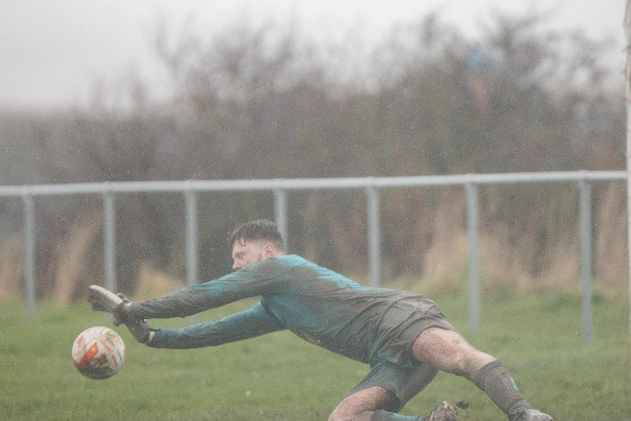 A soccer goalkeeper diving to save a ball during a game in rainy weather. The goalkeeper is wearing a teal and black uniform with gloves, and the field appears muddy.