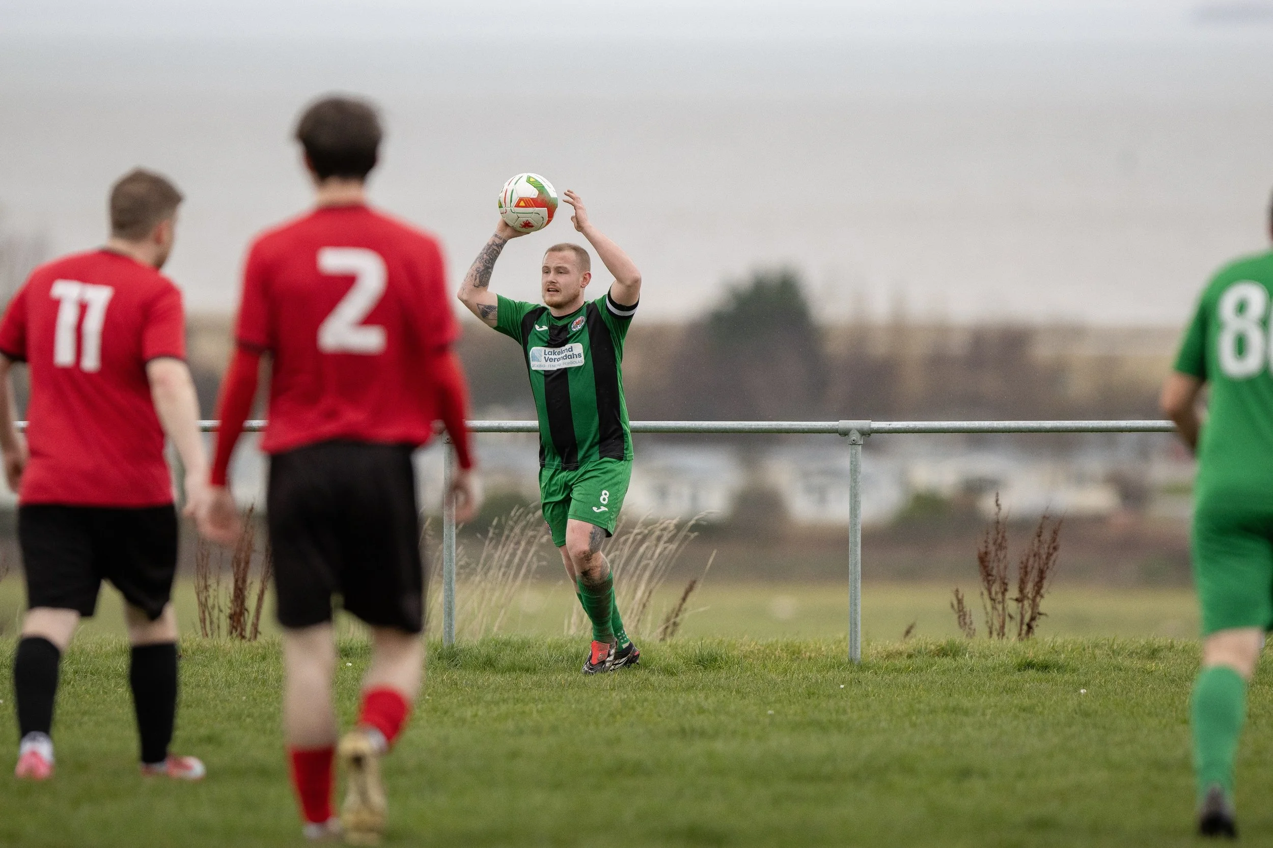 A soccer player in green and black uniform prepares to throw the ball in play during a match on a grassy field, with other players in red and green jerseys nearby.