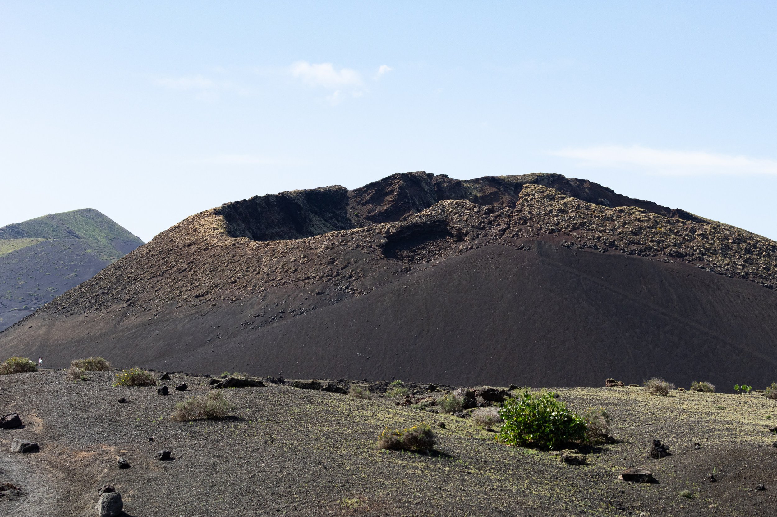Volcanic landscape with dark, rocky mountain and sparse desert vegetation under a partly cloudy sky.