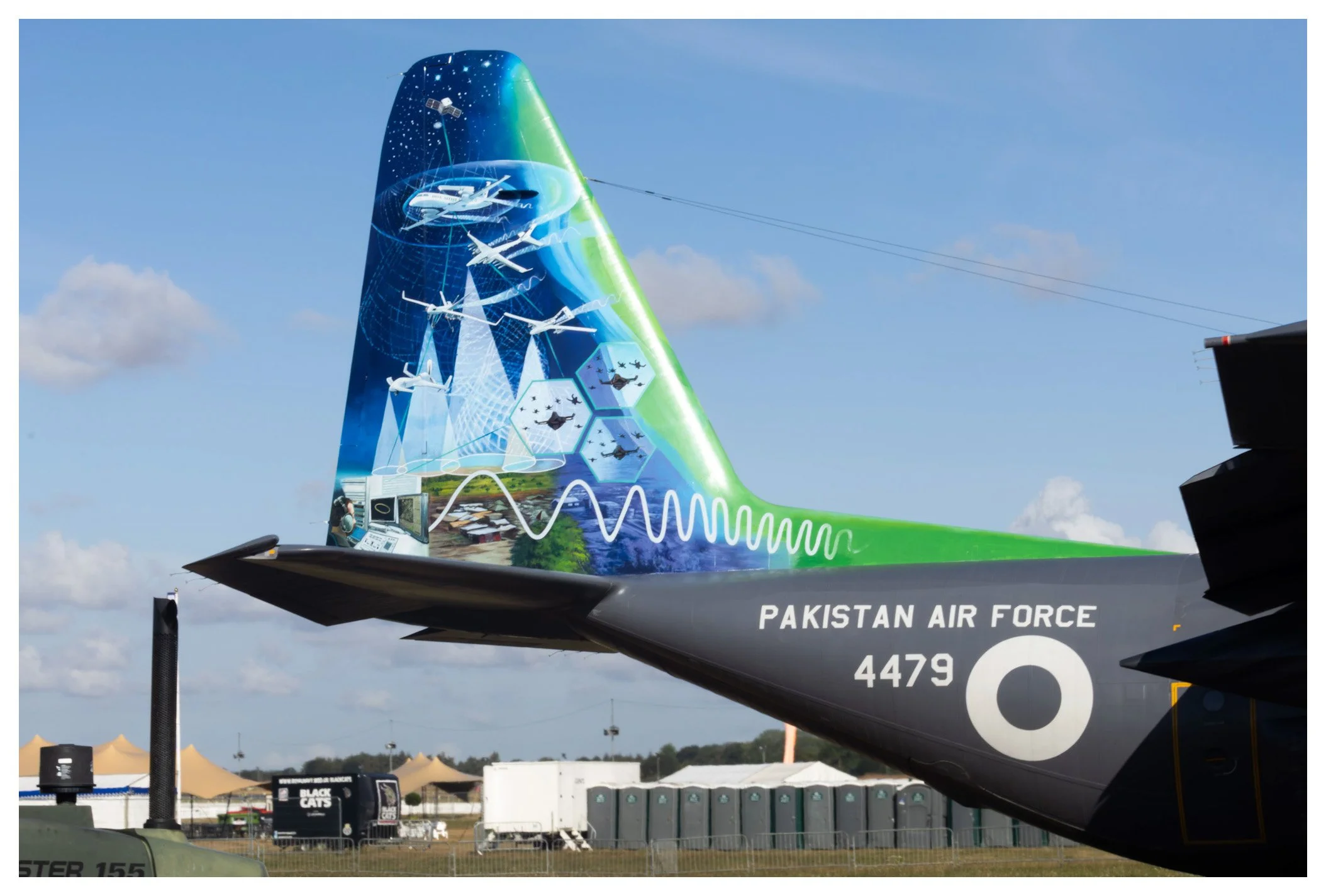 Pakistan Air Force jet with colorful artwork on its tail depicting airplanes, helicopters, and technology, with a clear sky and tents in the background.