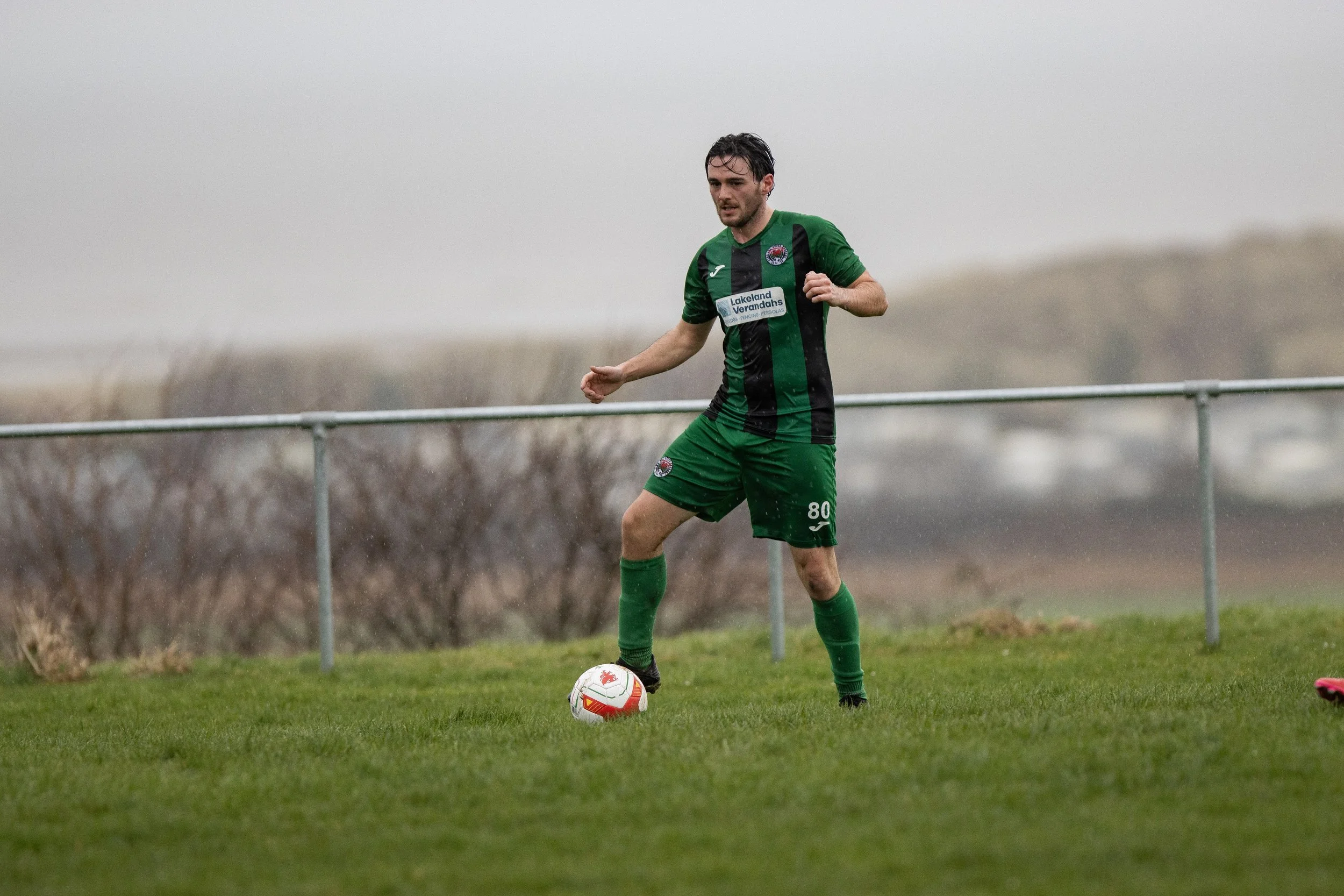 A soccer player in a green and black uniform with the number 80 is preparing to kick a soccer ball on a grassy field during a game in rainy weather.