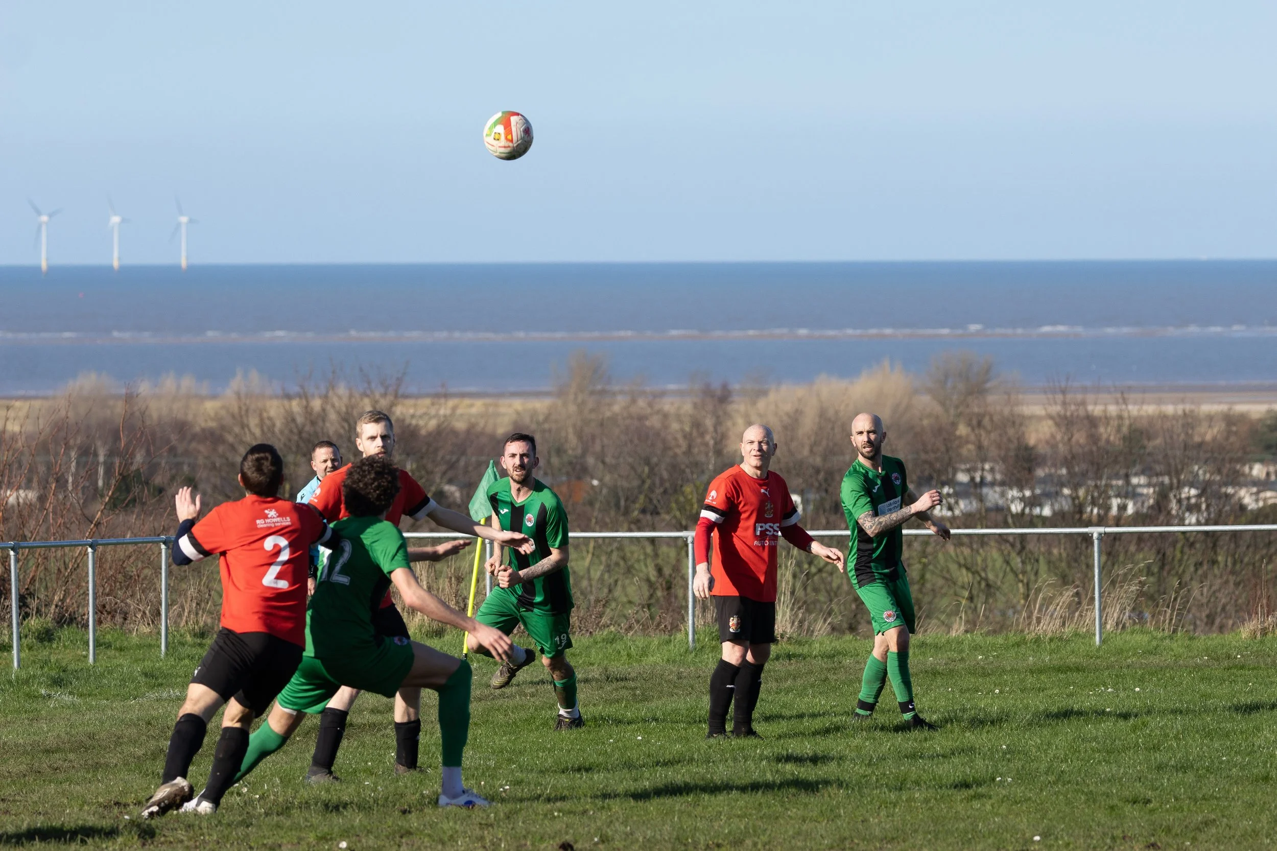 Soccer players in red and green jerseys playing on a grassy field near the coast, with wind turbines in the background, as a ball is mid-air above them.