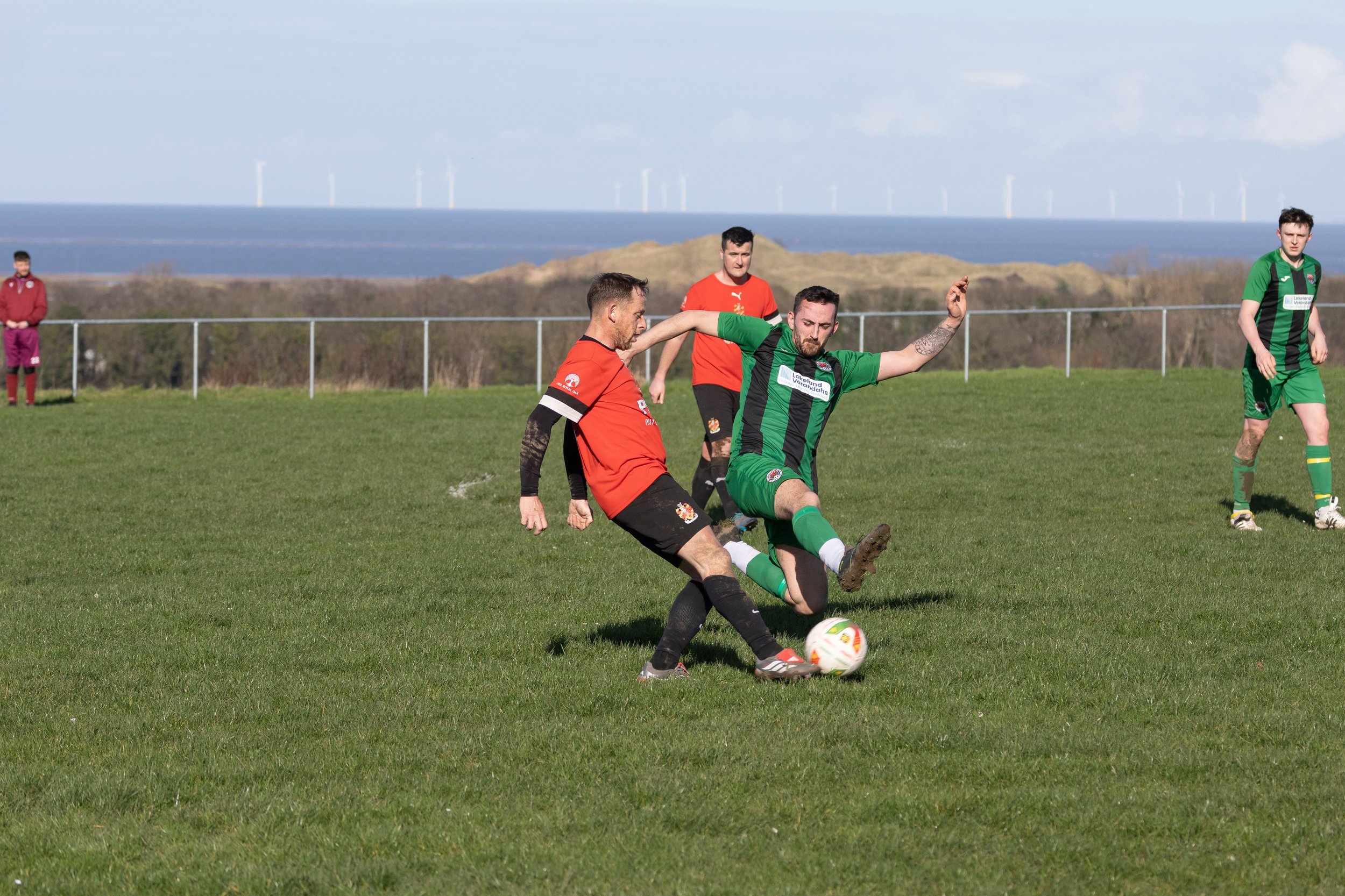 Soccer match with players in green and red jerseys competing for the ball on a grassy field, with a fence, shrubs, and a body of water with wind turbines in the background.