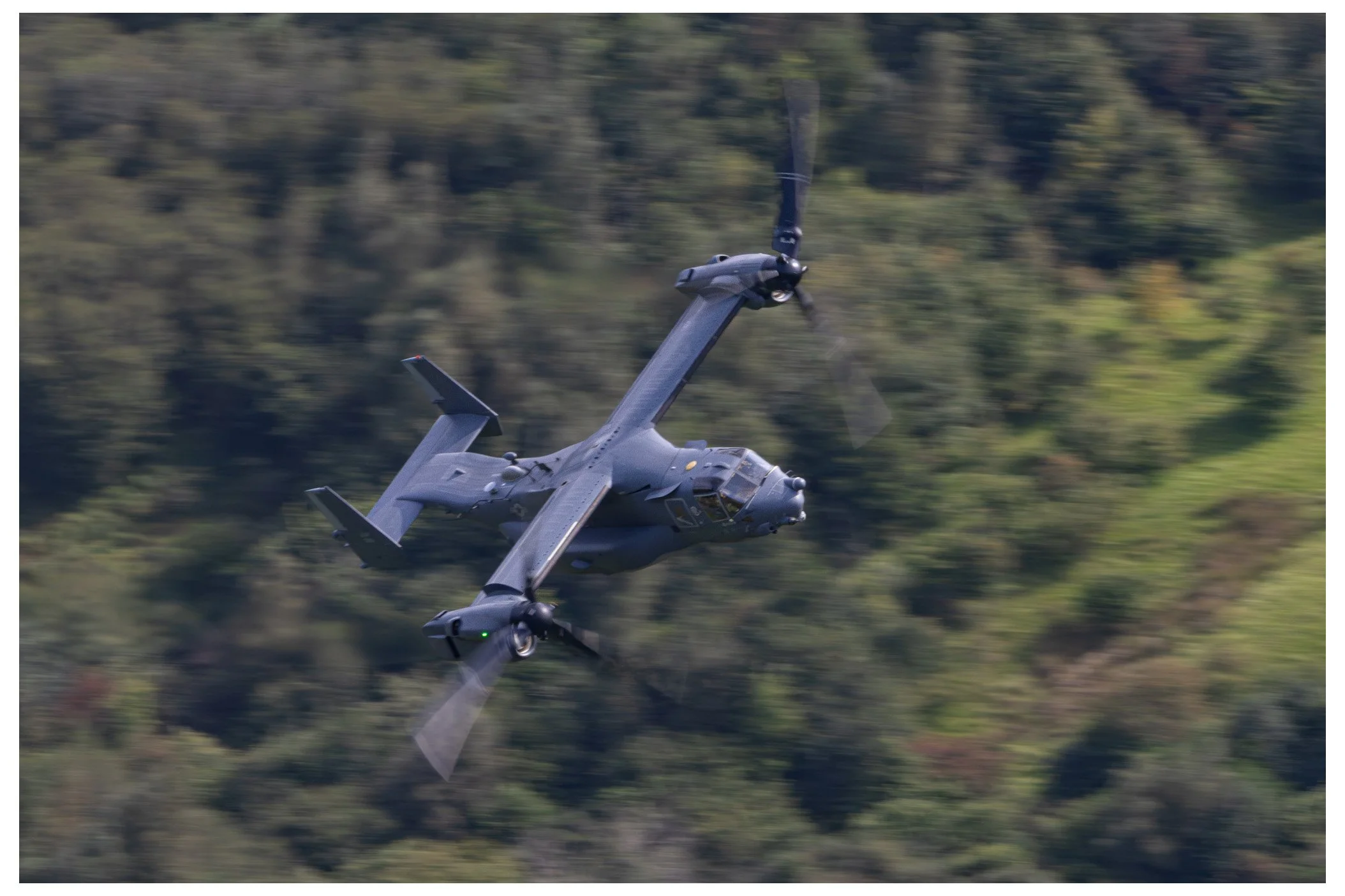 A military tiltrotor aircraft flying over a forested area, with its propellers spinning.