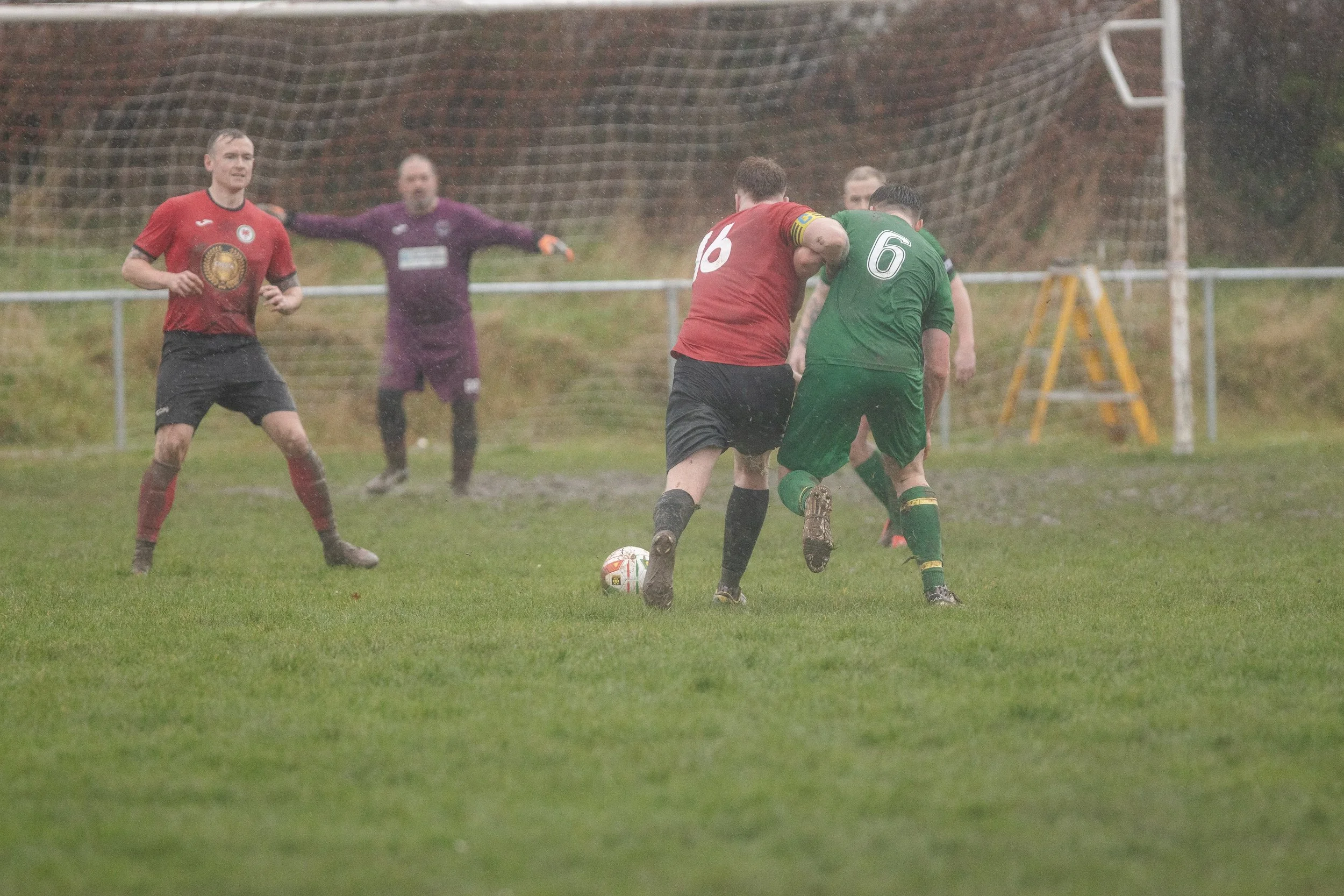 Two soccer players, one in a red jersey and one in a green jersey, contesting a ball on a rainy day on the field. Two other players, one in red and another in purple goalkeeper uniform, are nearby.