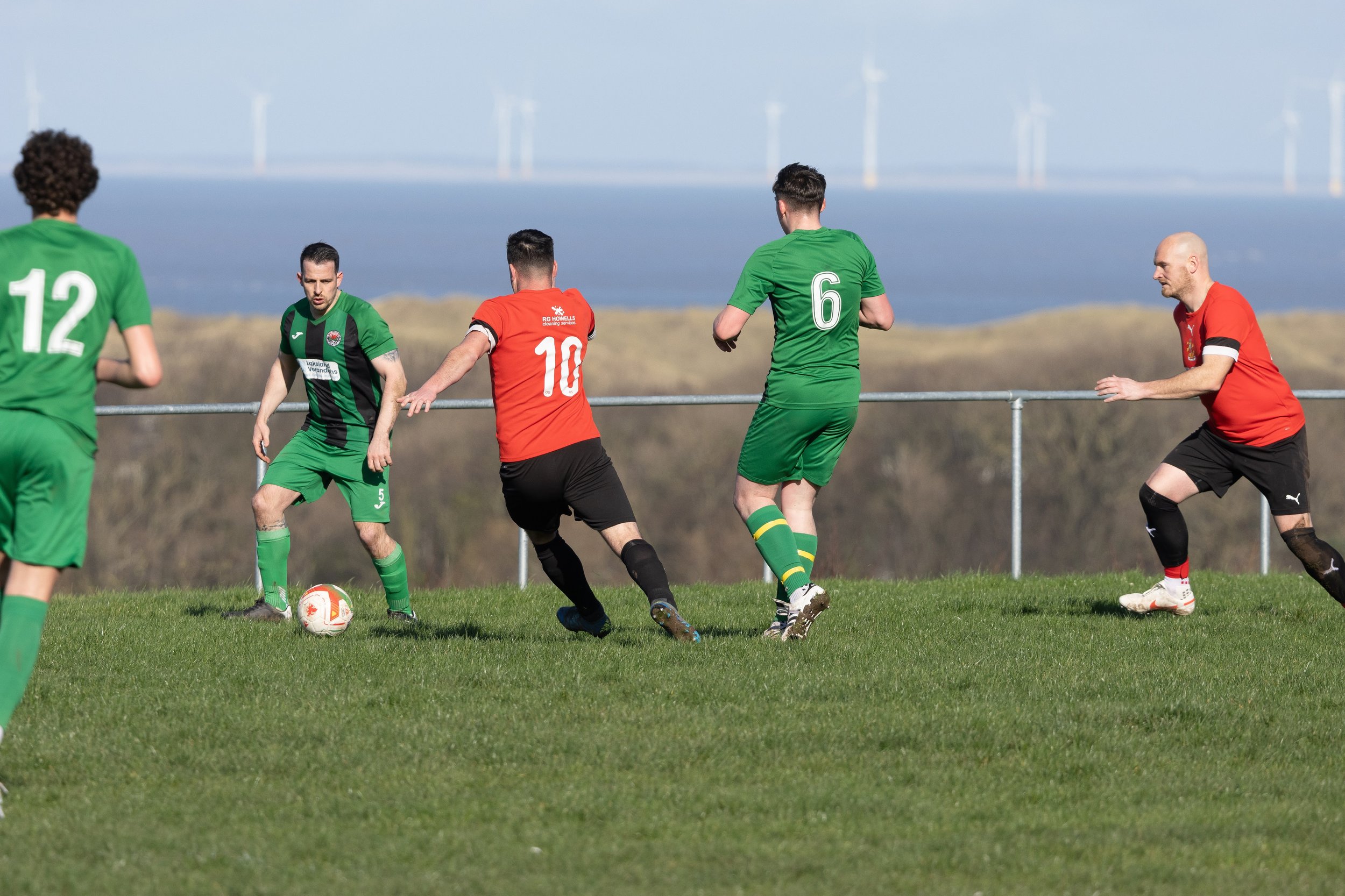 Soccer game with players in green and red uniforms playing on grass field, with a metal fence and wind turbines in the background.