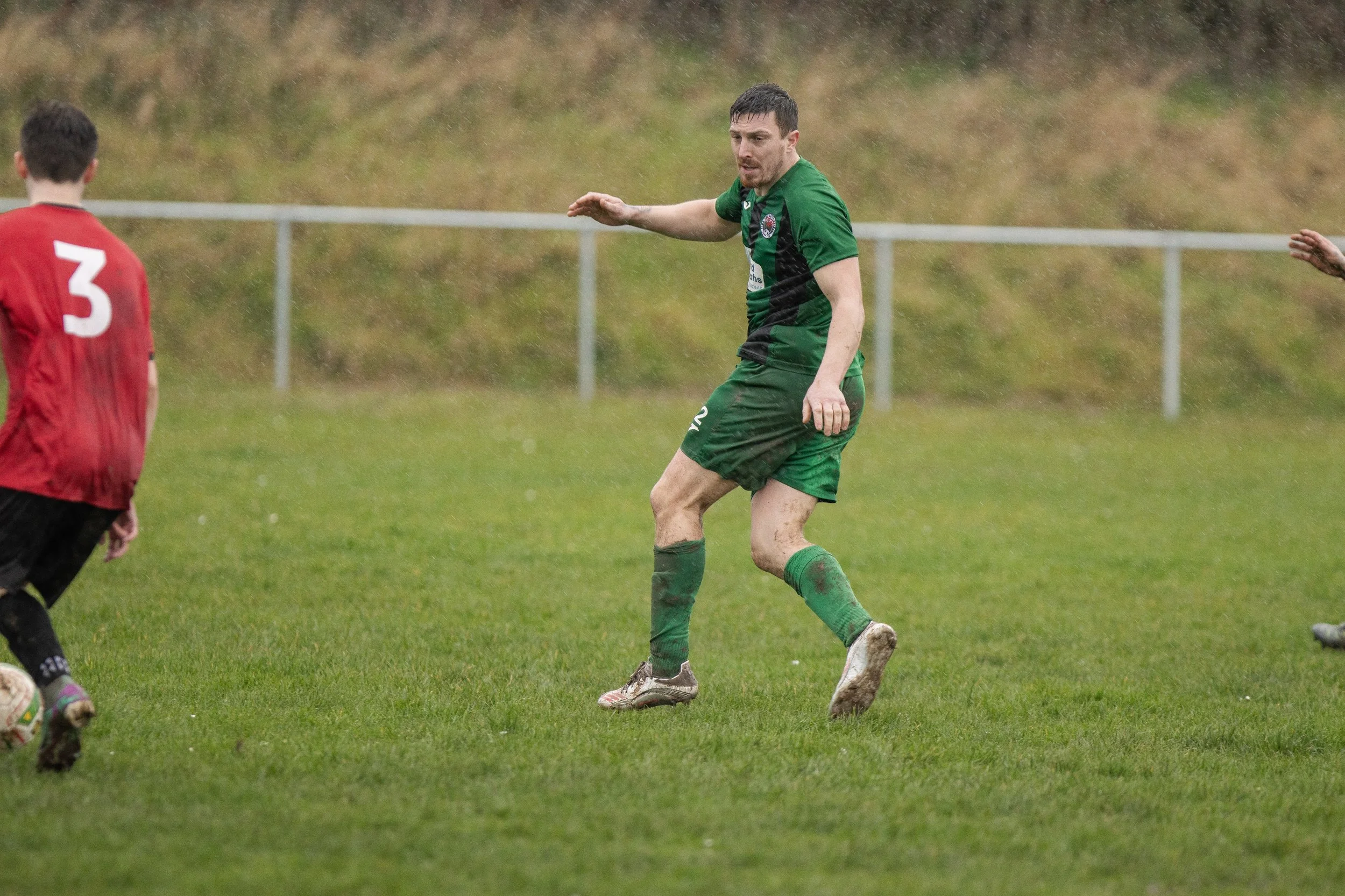 A soccer player in a green uniform on a rainy day, with muddy dirt on his legs, standing on a grass field during a match.