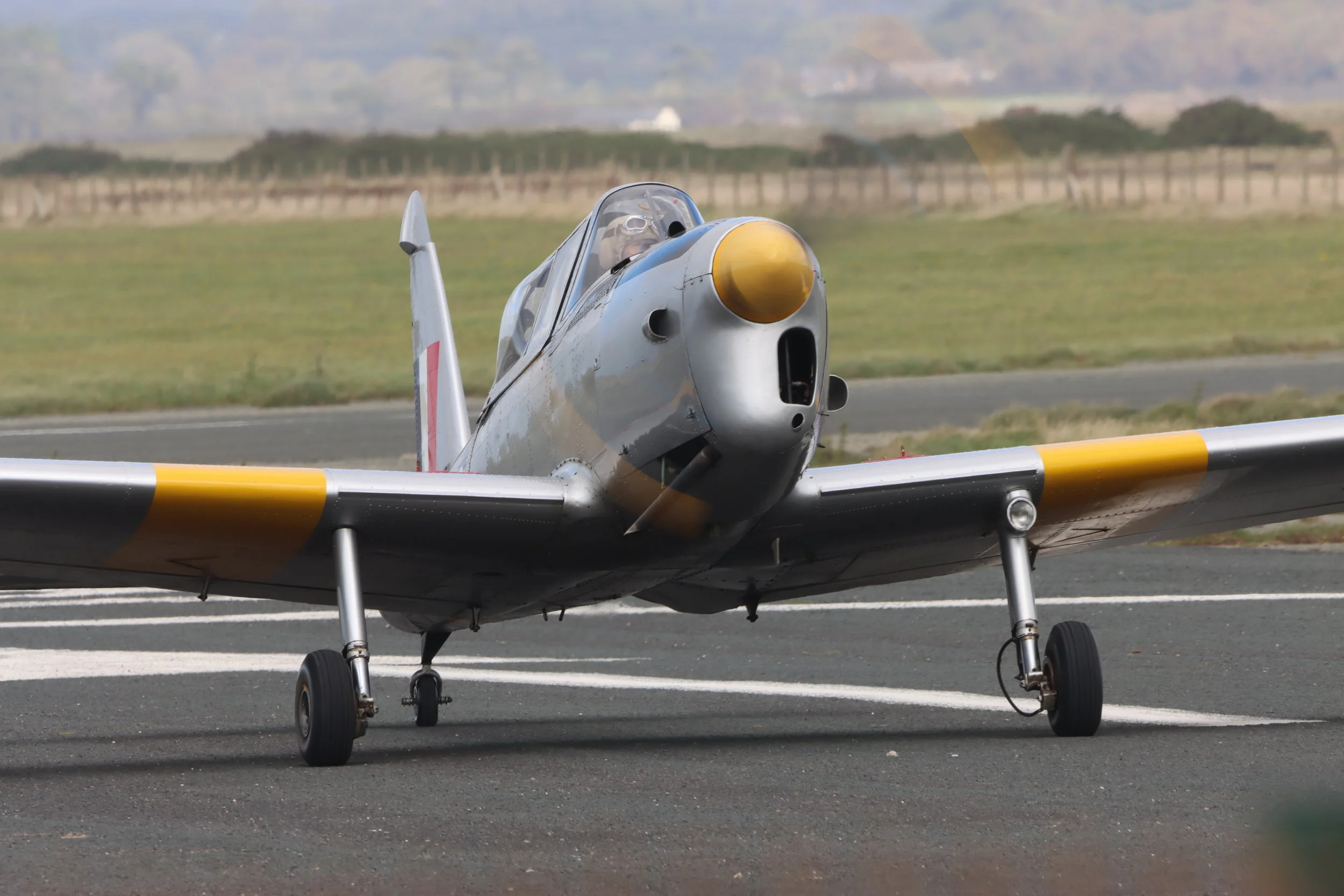 A vintage silver fighter jet with yellow accents on its nose and wingtips parked on a runway during the daytime.
