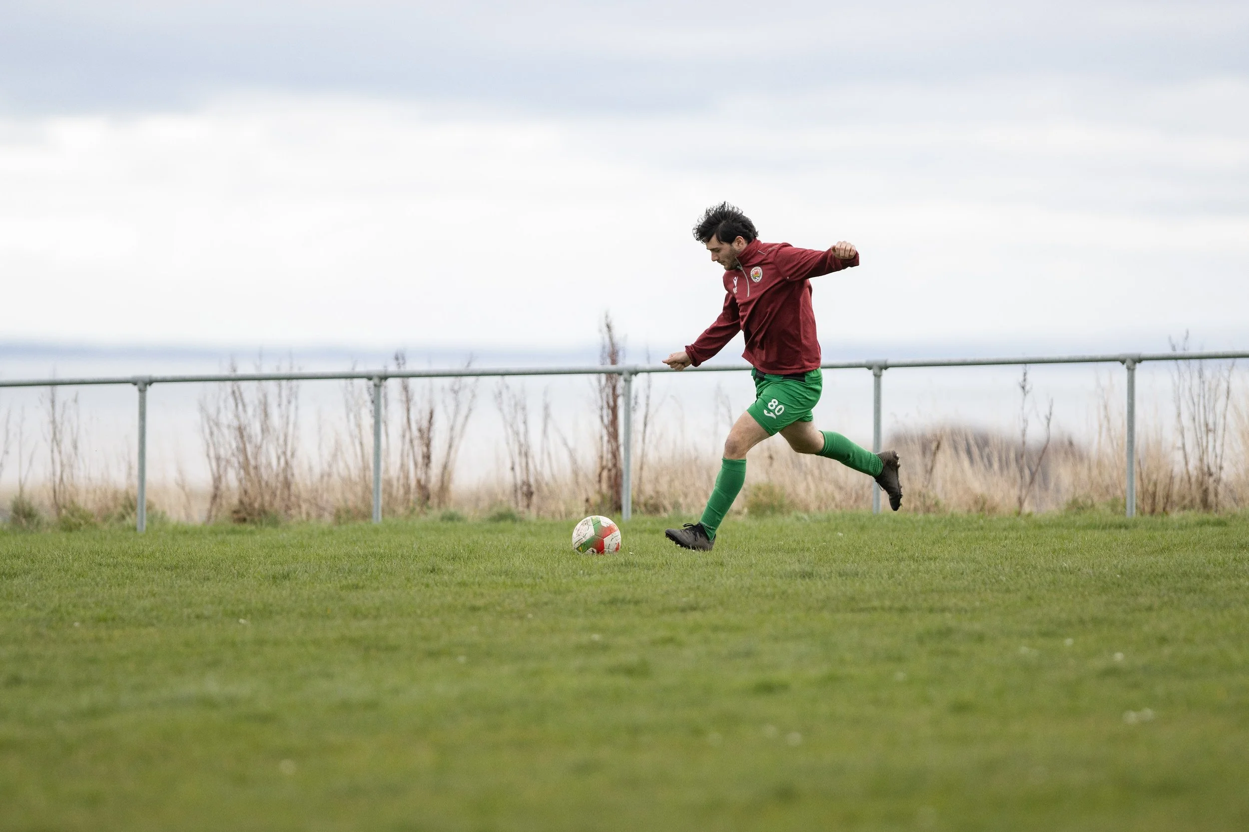 A man in a maroon sports jacket and green shorts playing soccer on a grassy field.