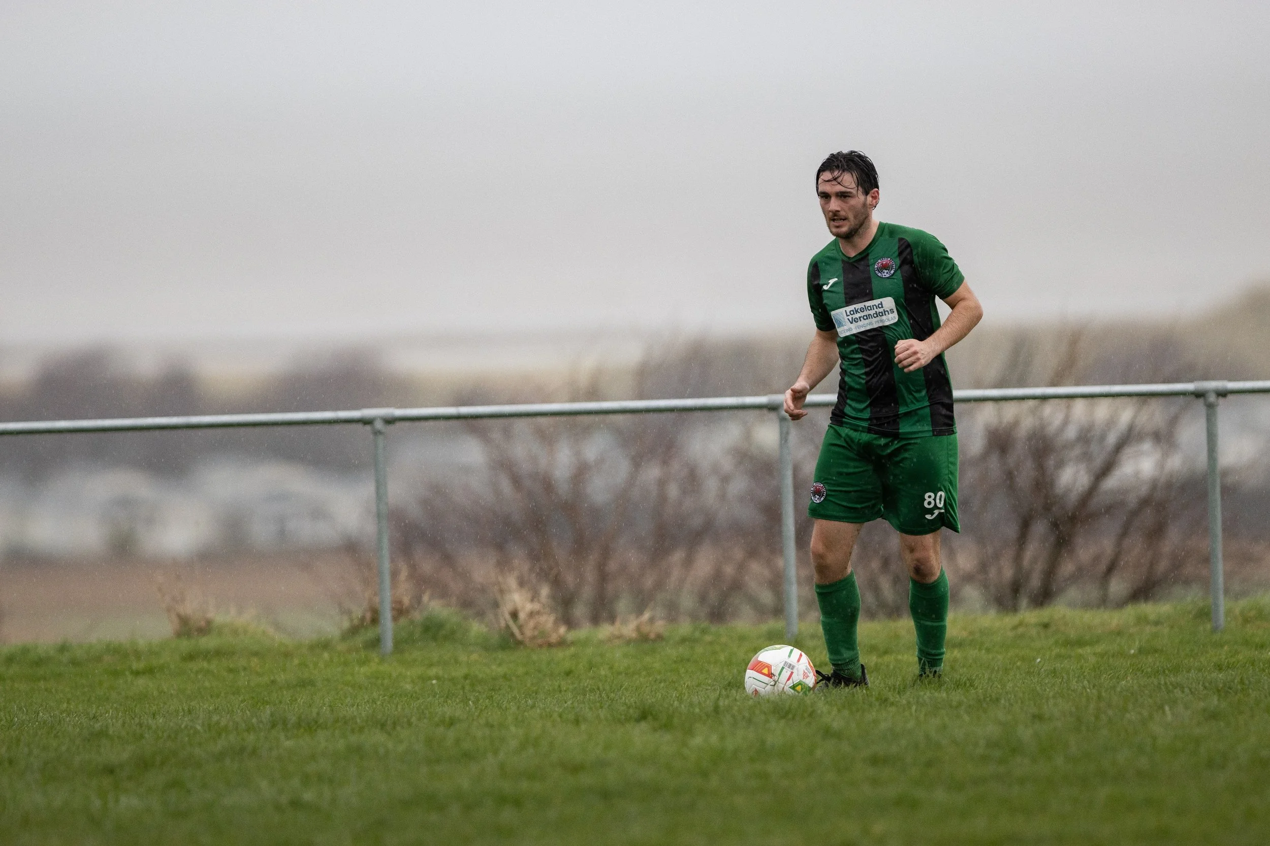 A male soccer player in a green and black uniform preparing to kick a soccer ball on a grassy field on a rainy day.