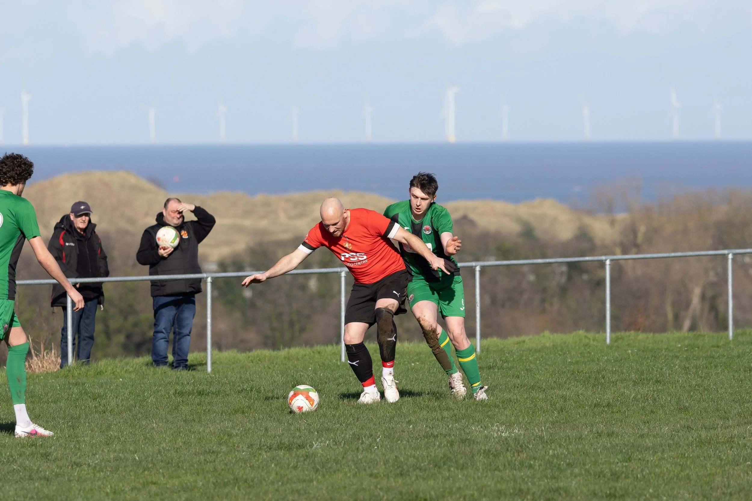 Two soccer players competing for the ball on a grassy field, with a scenic background of hills, trees, and wind turbines, and spectators watching nearby.