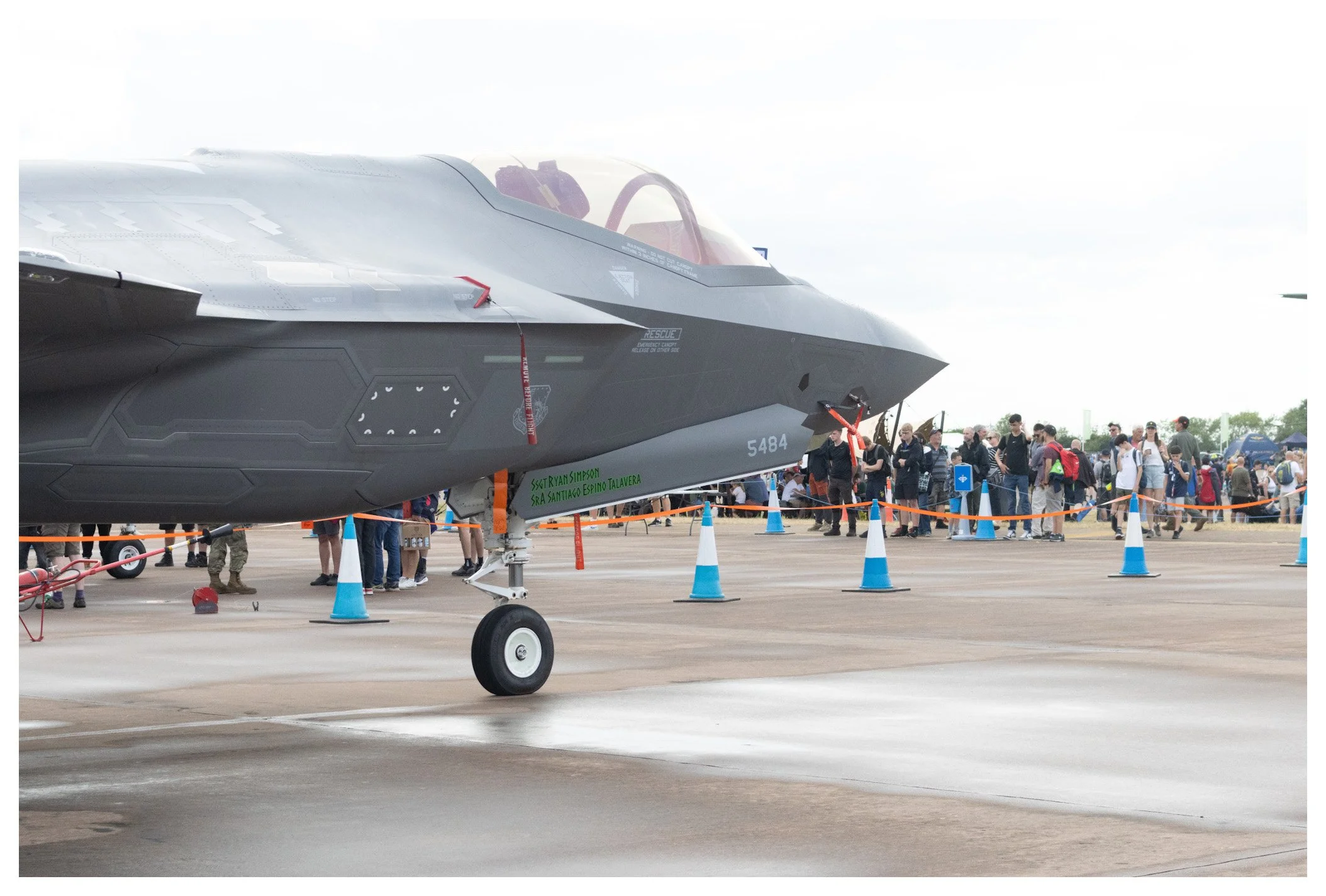 A military fighter jet on display at an air show with a crowd of people in the background, surrounded by orange and blue safety cones.